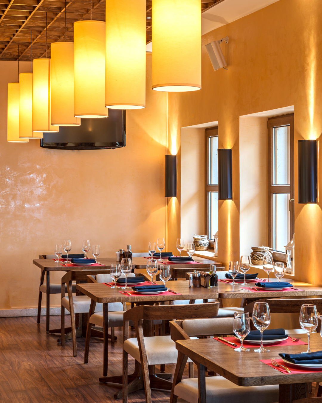 Interior view of a restaurant with wooden tables set with wine glasses, black napkins, and red placemats illuminated by large hanging yellow lanterns and wall-mounted lights, near windows on a yellow wall.