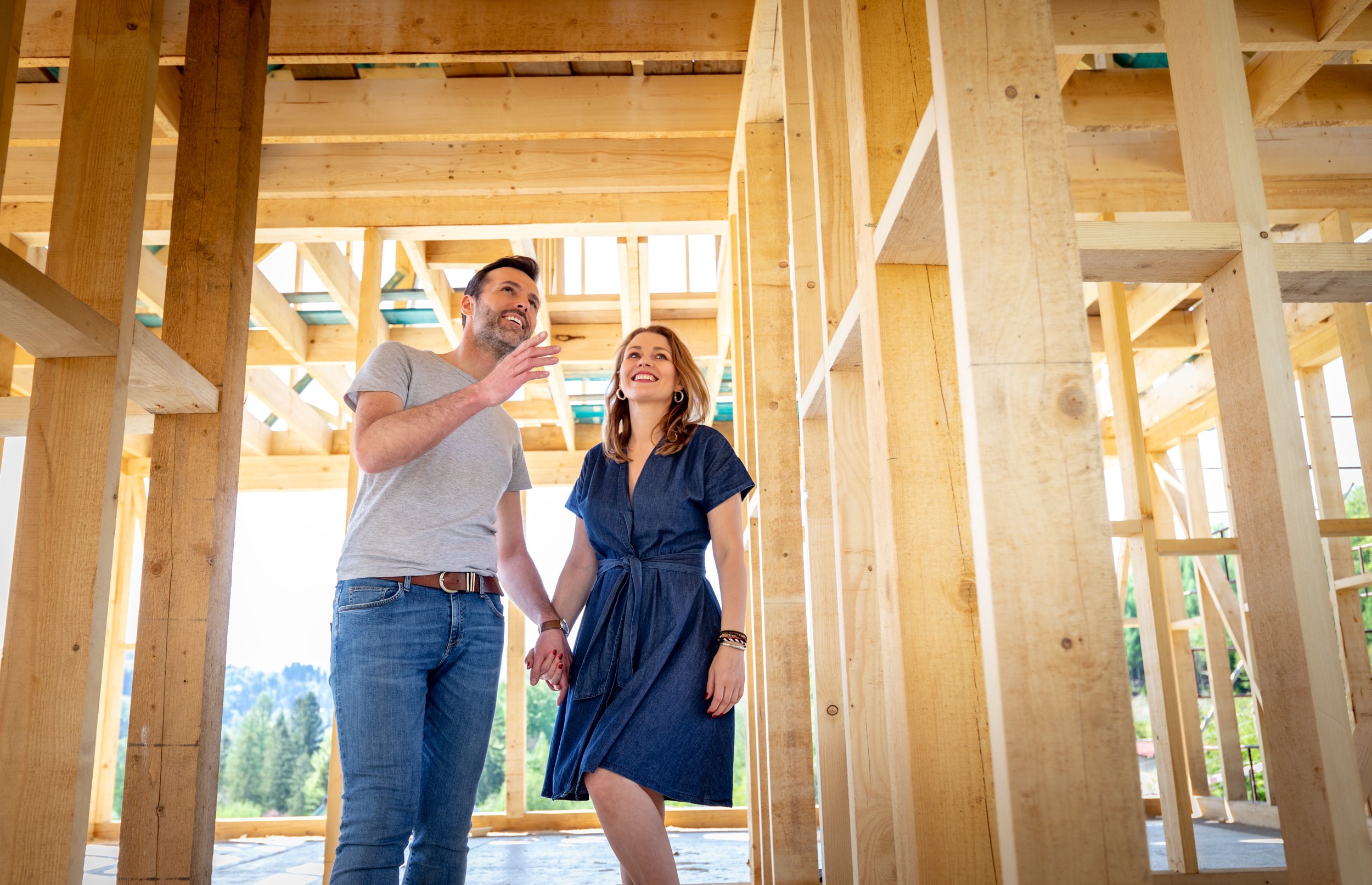 A couple standing inside a wooden house under construction, holding hands, smiling, and talking to each other.