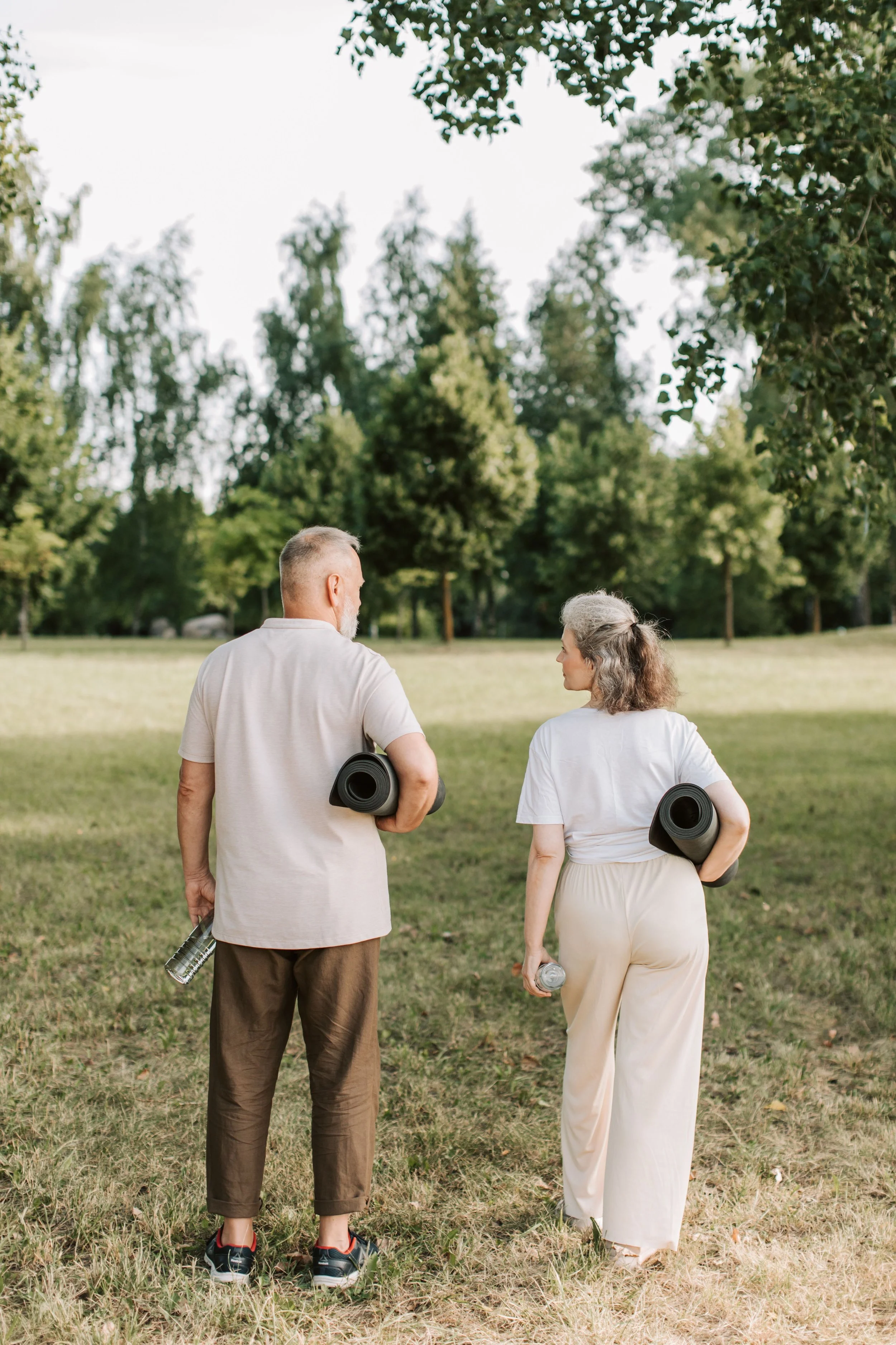 Una parella de persones adultes caminant per un parc amb estoretes d’exercici sota el braç i una ampolla d’aigua a la mà. La imatge representa un estil de vida actiu, l’exercici físic a l’aire lliure i la promoció de la salut i el benestar.