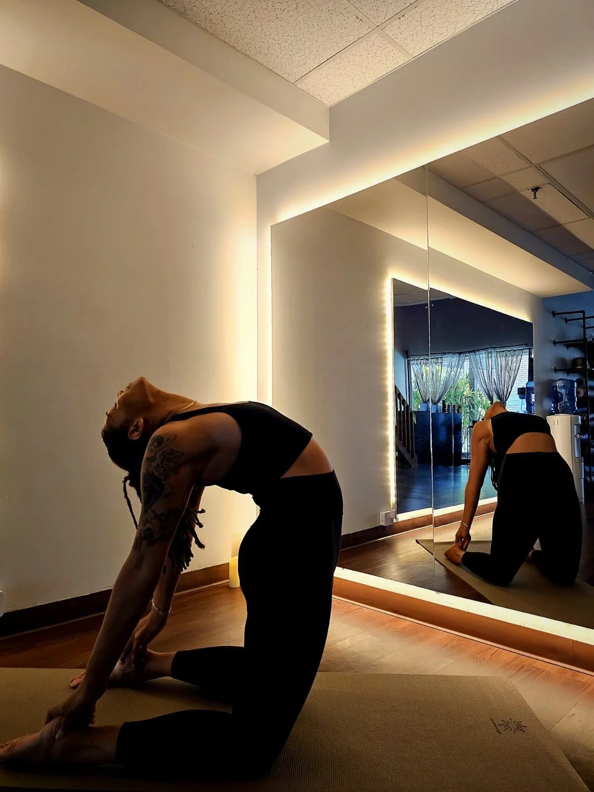 A woman with curly hair performing a yoga pose indoors, sitting on a colorful rug against a wall with abstract decorative patterns. She is wearing a yellow top and olive-green pants, and is practicing in front of a projected or painted background.