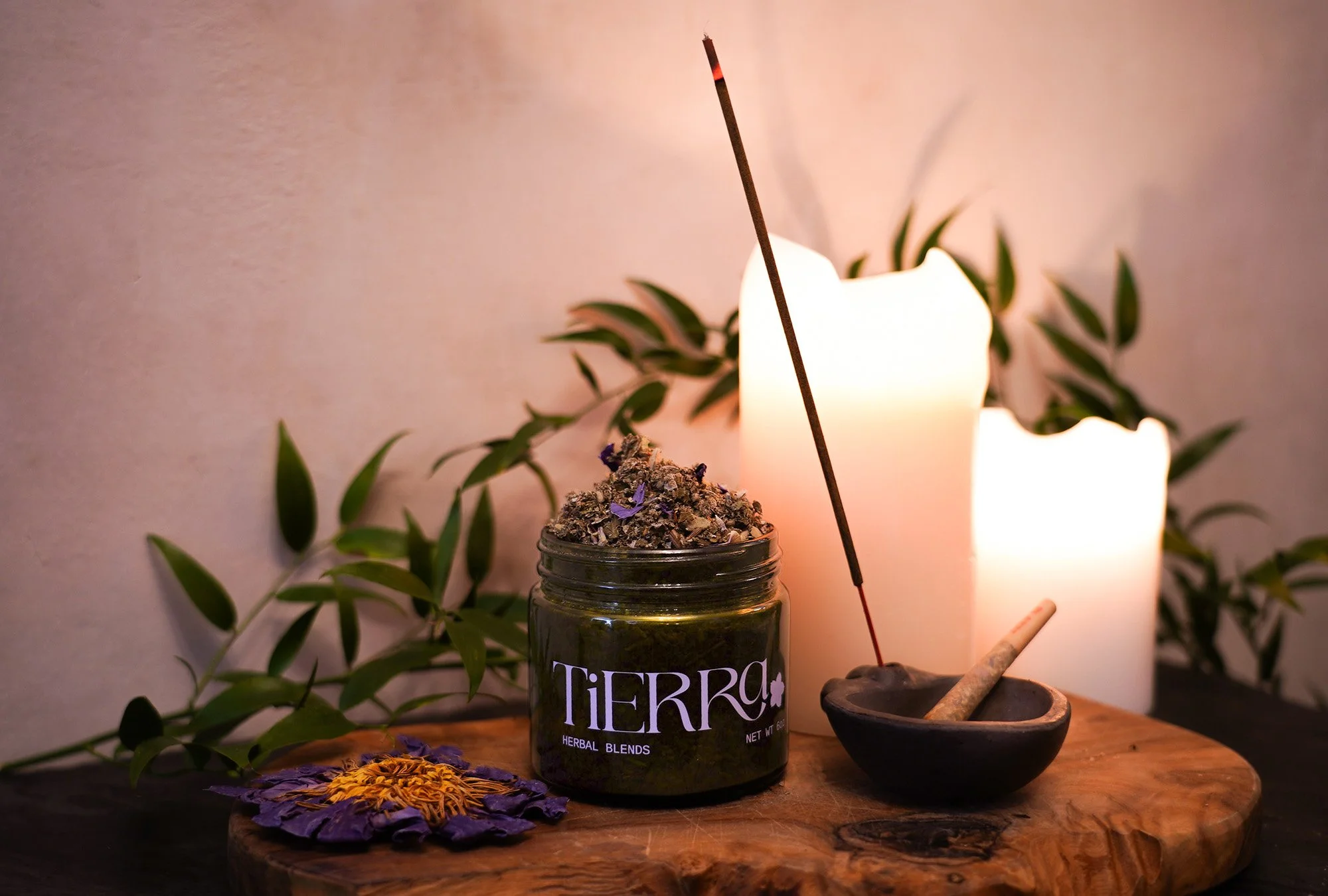 A jar labeled 'TIERRA Herbal Blends' with dried herbs on top, a lit incense stick in a black bowl, two white candles, and a purple flower on a wooden surface with green leafy background.