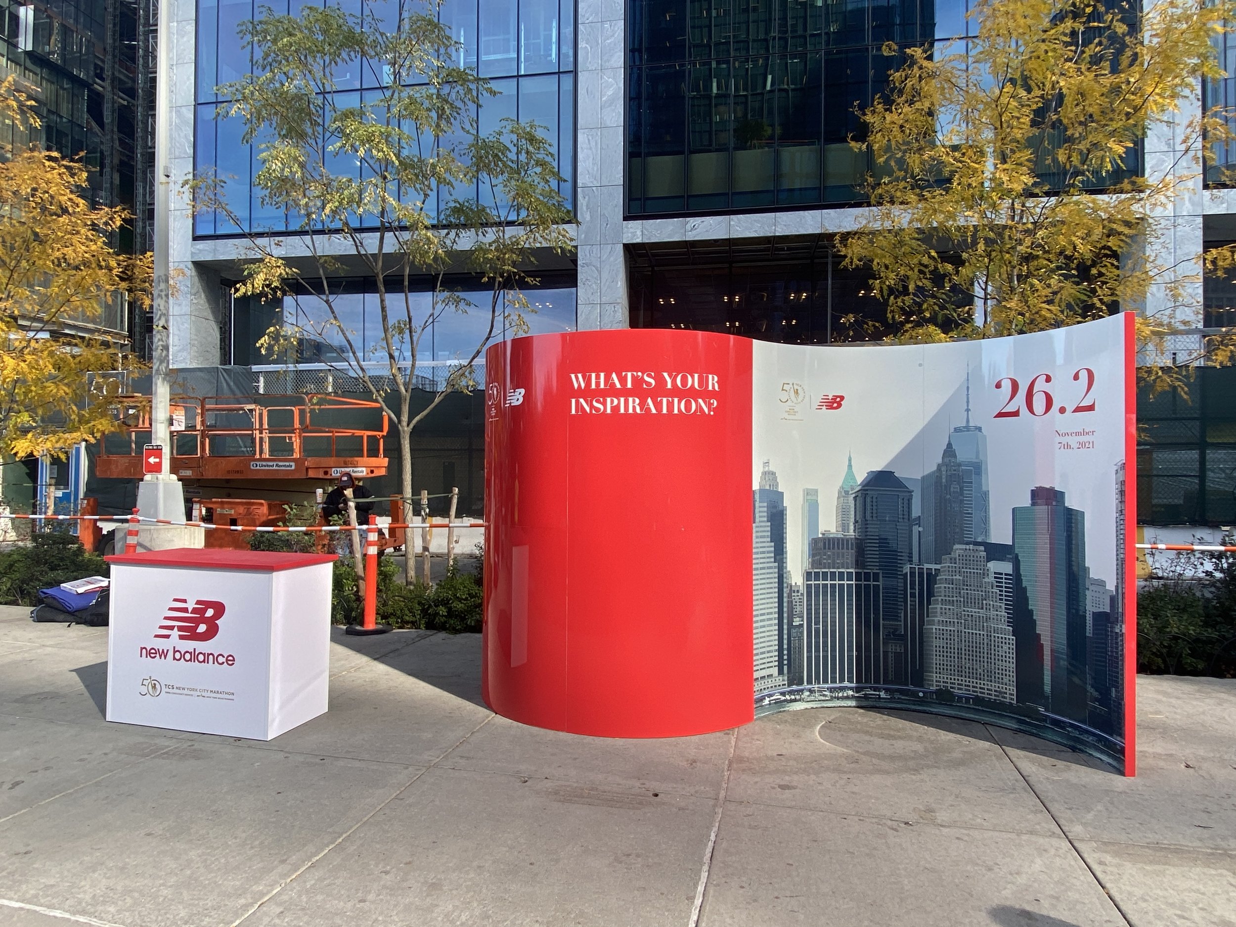 Outdoor display featuring New Balance branding and promotional material for the New York City Marathon, with a city skyline backdrop and trees with yellow leaves.
