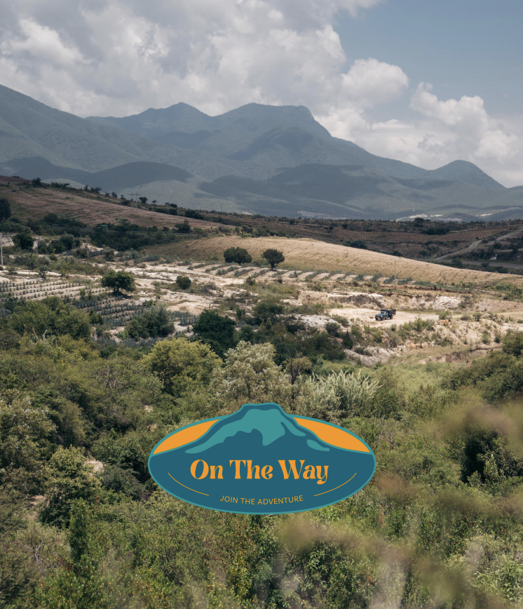 A landscape with mountains in the background, rolling hills, and green vegetation in the foreground. There is a vehicle on the road and a logo with a mountain and the text 'On The Way' and 'Join the Adventure' at the bottom center.
