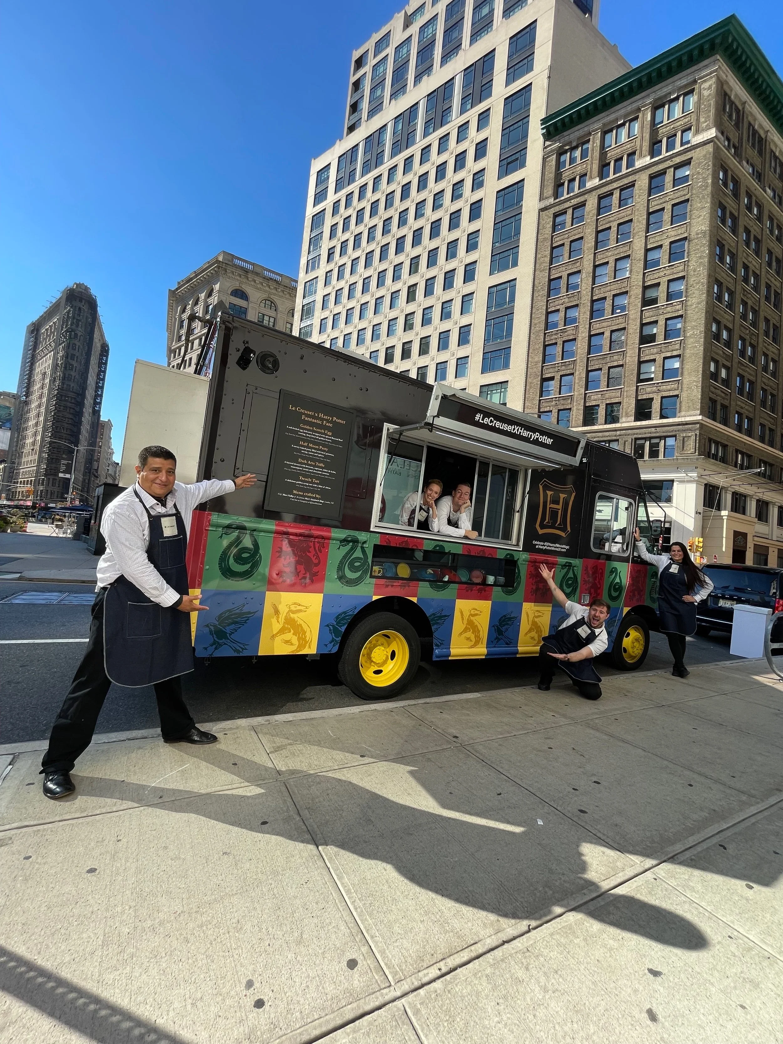 Four people dressed as Harry Potter characters posing next to a colorful Harry Potter-themed food truck on a city street with tall buildings in the background.