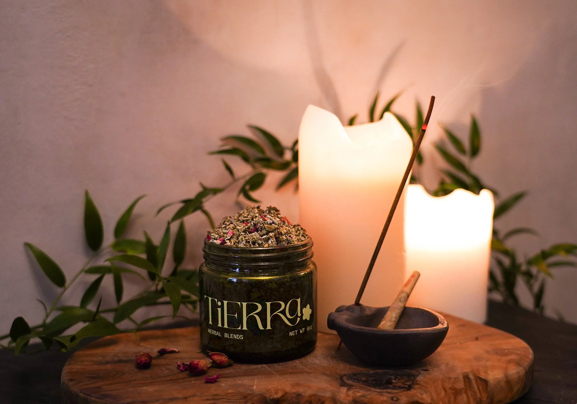 A wooden table with a jar of herbal blend labeled 'TiERRA', a small black bowl with incense and a stick of incense, and two lit candles in the background, with green leafy plants.