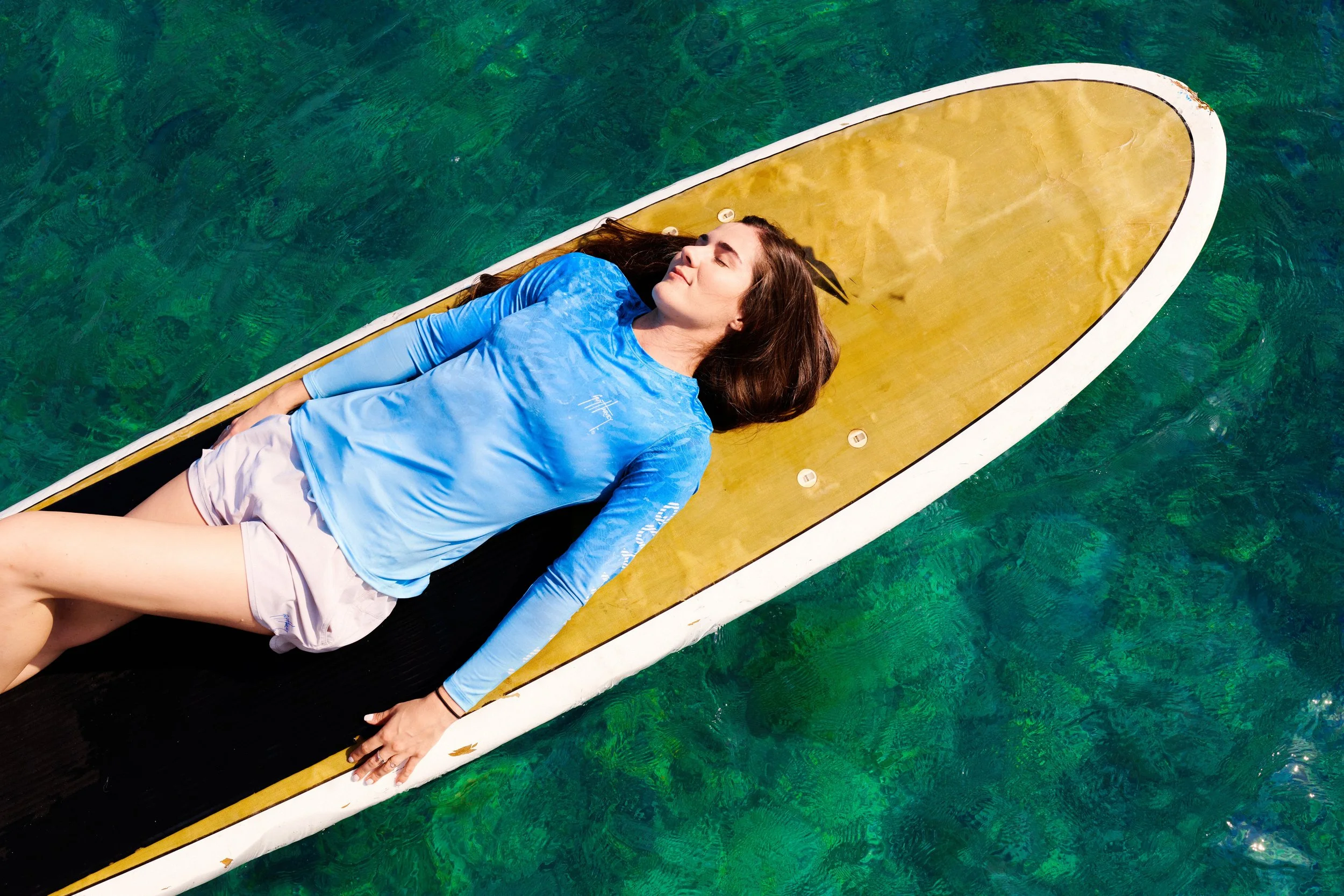 A woman lying on her back on a paddleboard in clear green water, wearing a blue long-sleeve shirt and light-colored shorts, with her eyes closed and face relaxed.