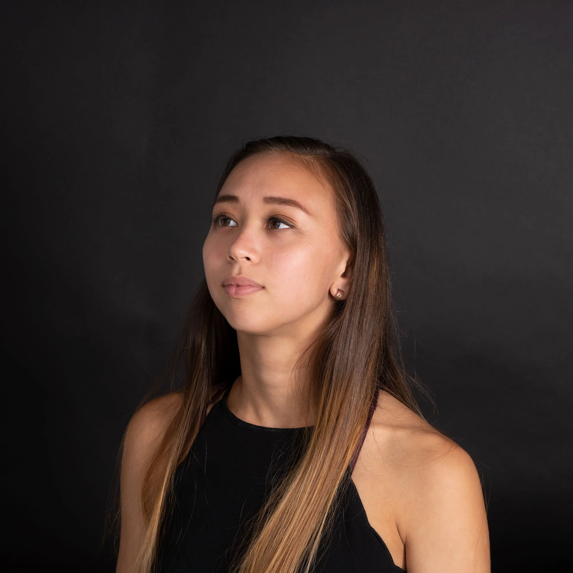 Portrait of a young woman with long brown hair wearing a black sleeveless top, looking slightly to the side against a dark background.