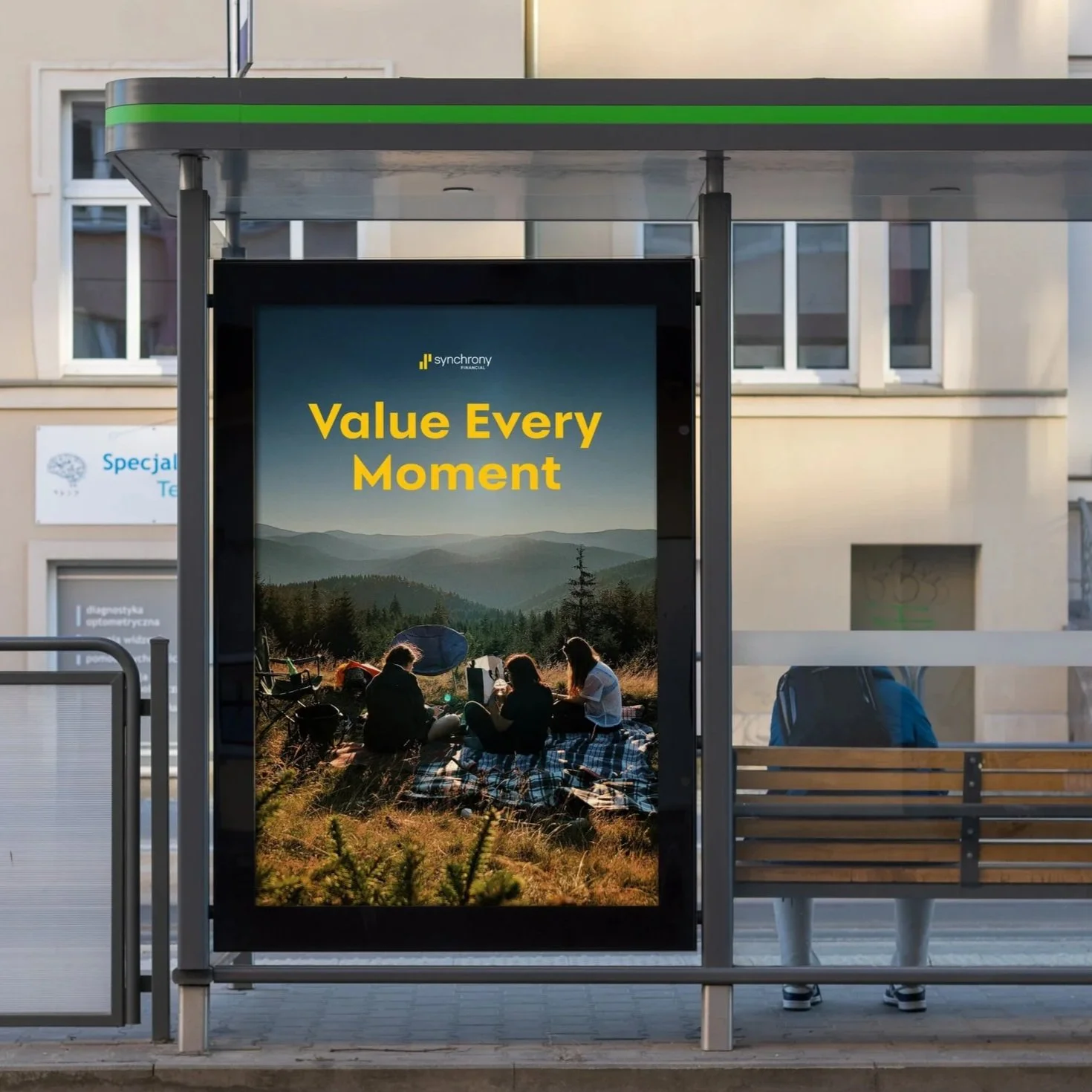 Bus stop shelter with an advertisement featuring a landscape with mountains and trees, and a group of people sitting on a grassy hill having a picnic at sunset, with the text 'Value Every Moment' in yellow.