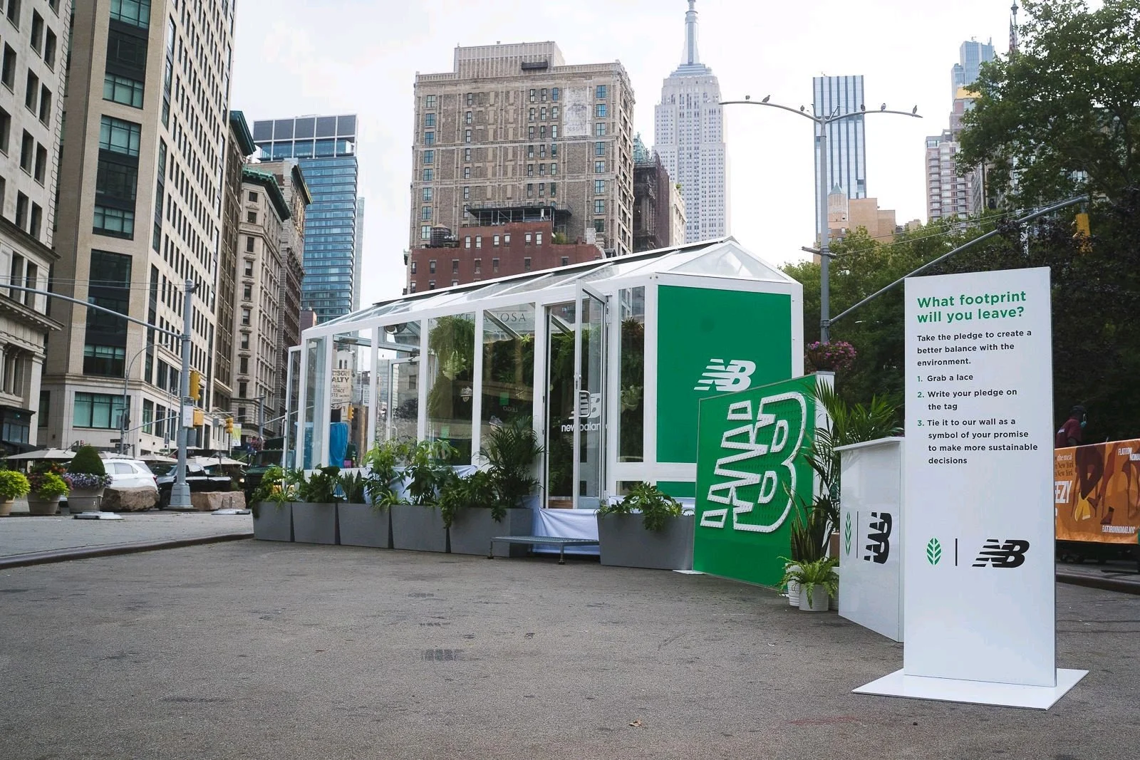 An outdoor eco-friendly booth with plant decorations and New Balance branding, featuring a sign with environmental pledge steps in a city street with tall skyscrapers in the background.