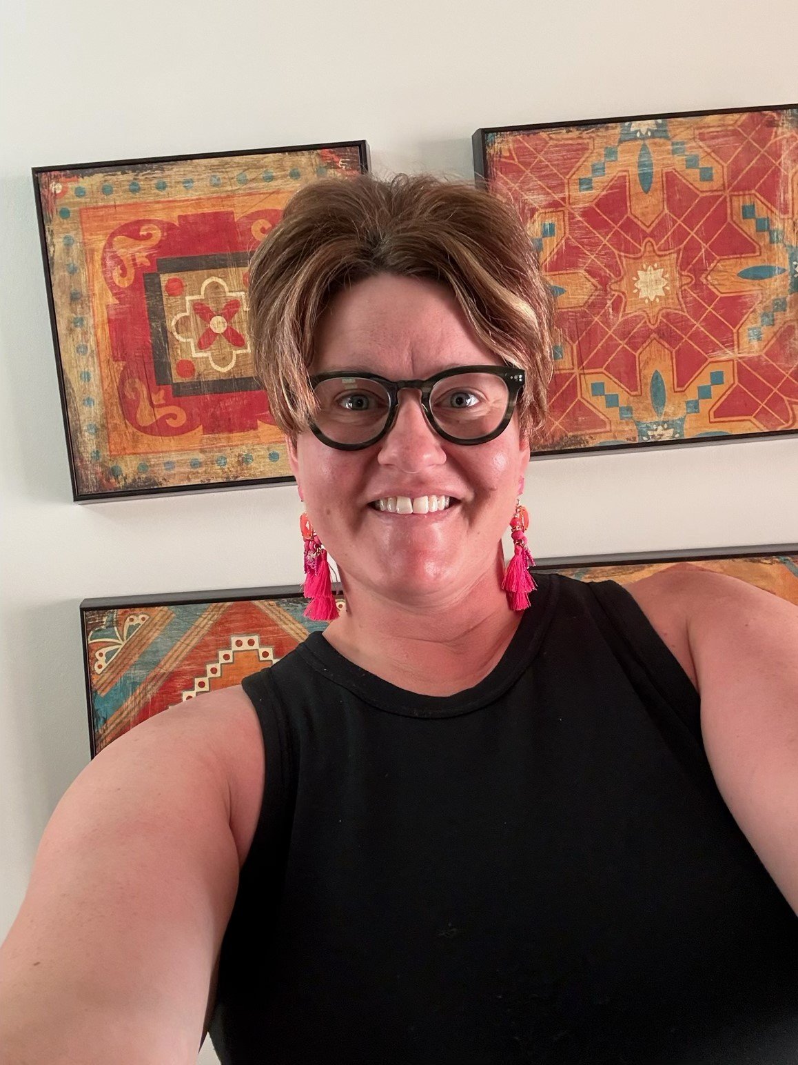 A woman with short brown hair, wearing black glasses, pink earrings, and a black sleeveless top, smiling for a selfie in front of colorful framed artwork.