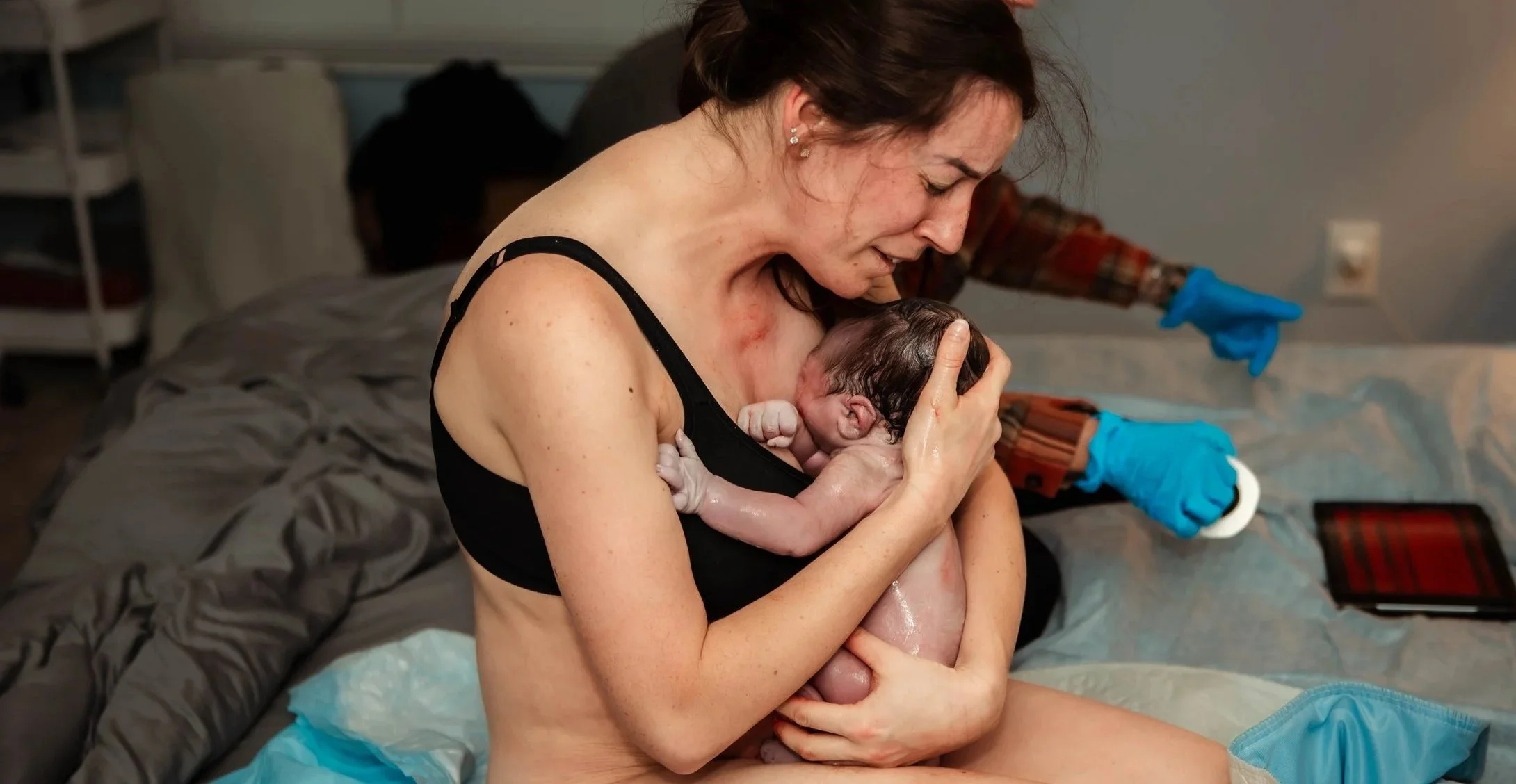 A woman holding a newborn baby during childbirth in a hospital room, with medical staff in the background.