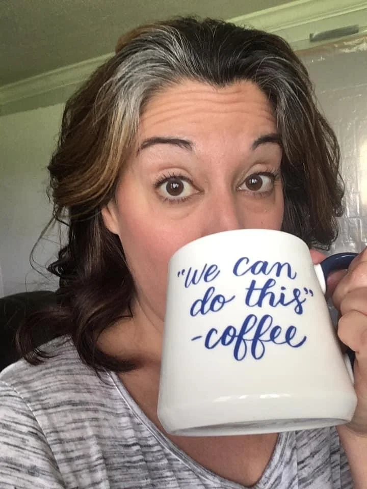 A woman with brown and gray hair holding a white coffee mug in front of her face. The mug has the quote 'we can do this' - coffee written on it in blue text.