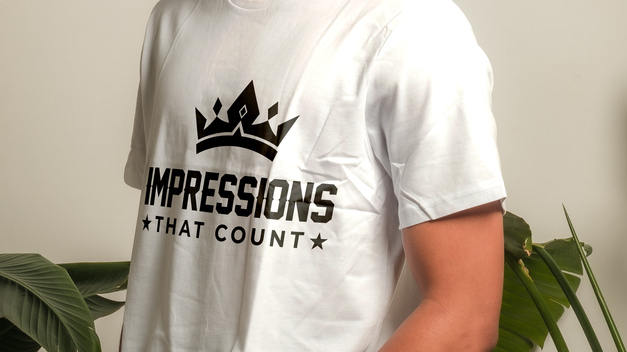 Person wearing a white T-shirt with a black graphic of a crown and the text 'IMPRESSIONS THAT COUNT', standing next to green leafy plants.