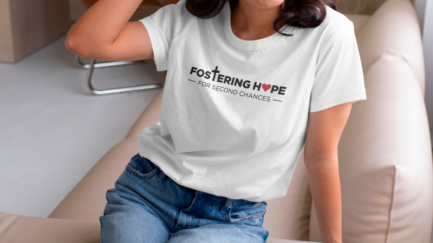 Person wearing a white T-shirt with fostering hope text and a red heart symbol, sitting on a beige couch.