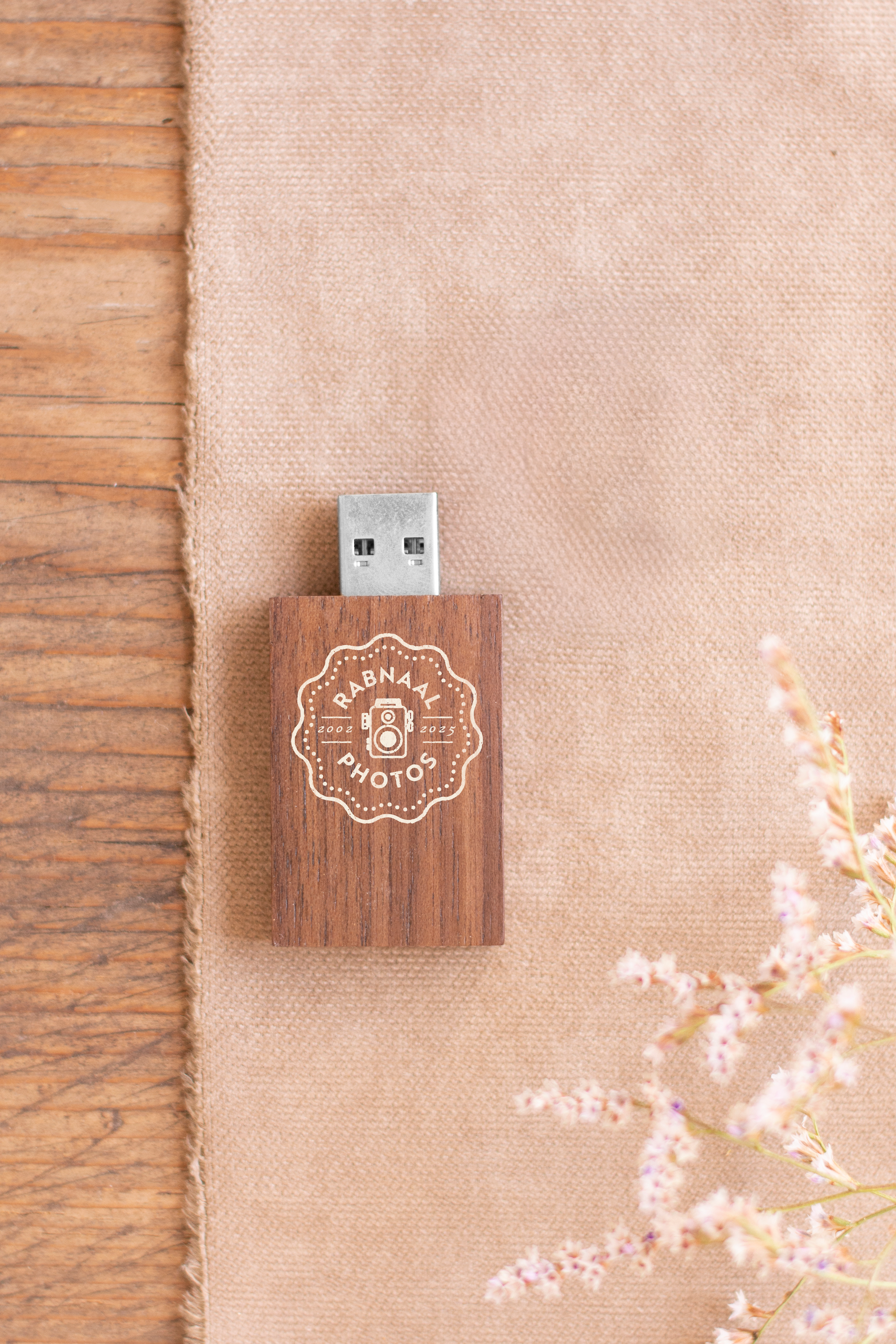 A USB flash drive with a wooden case and a logo that reads 'Rabnaal Photos 2002-2025' along with a camera icon, on a beige fabric surface next to a wooden surface and some pink flowers.