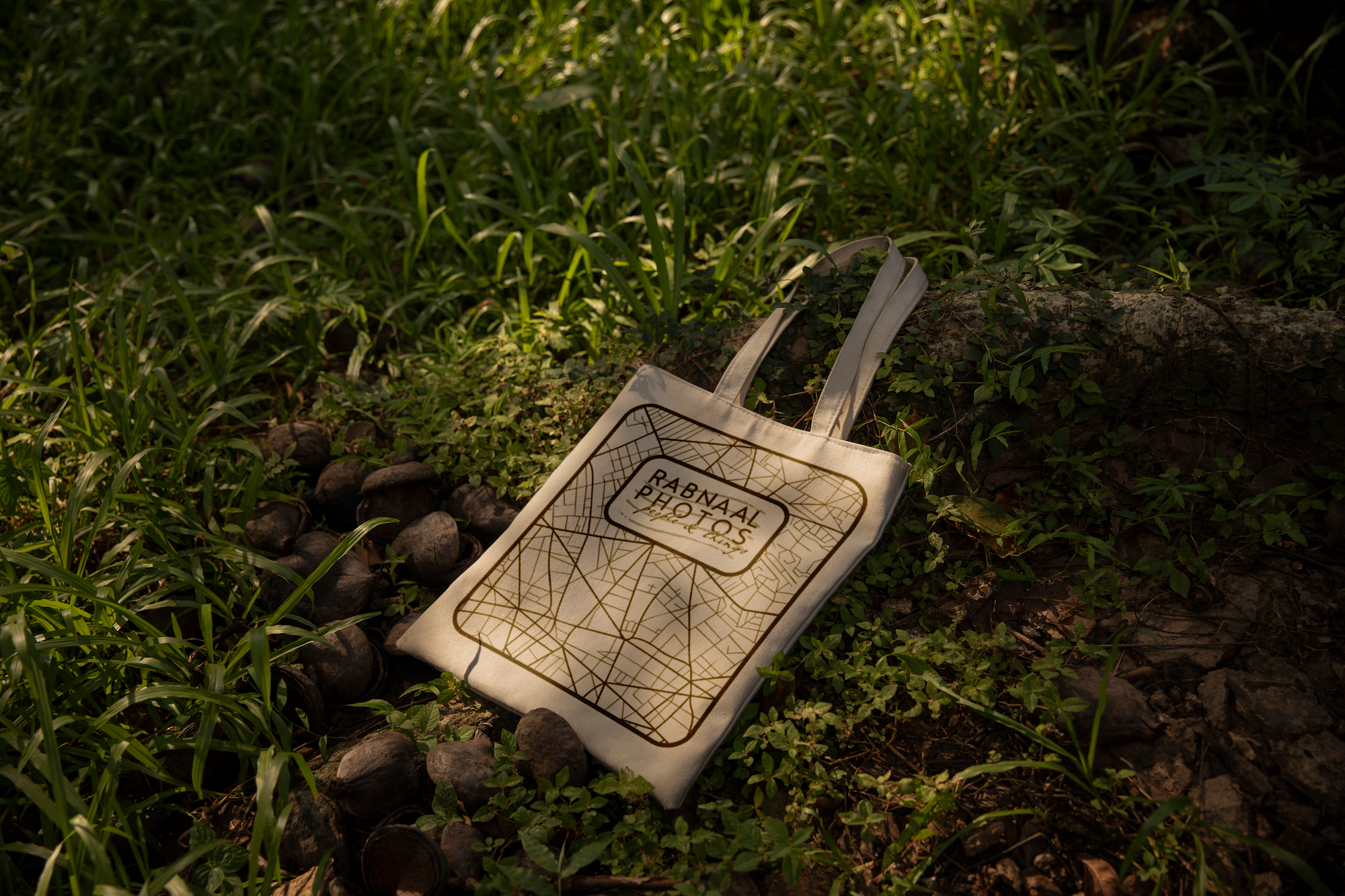 A tote bag with a city map and the text 'RABNAAL PHOTOS' rests on the ground among grass and small rocks in a natural setting.