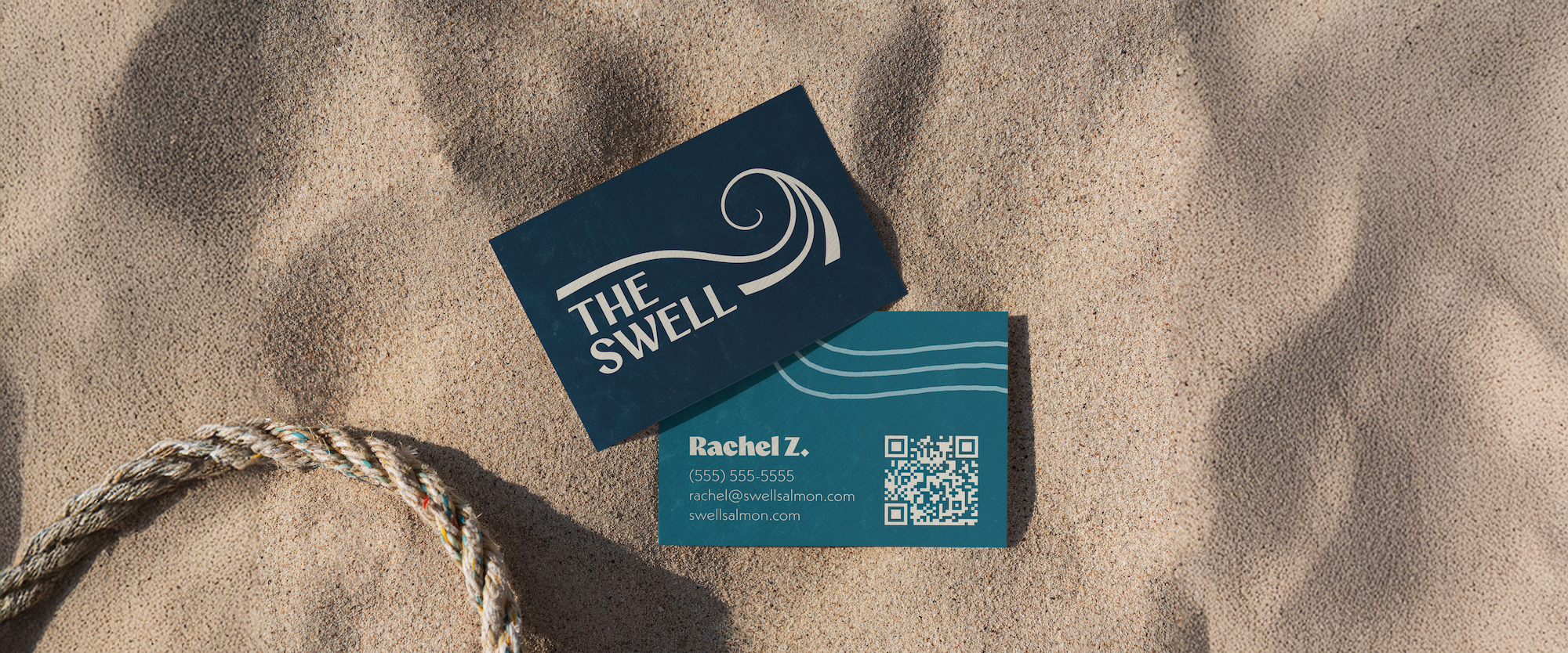 A business card and a sticker on sandy beach with a rope, related to 'The Swell' branding.