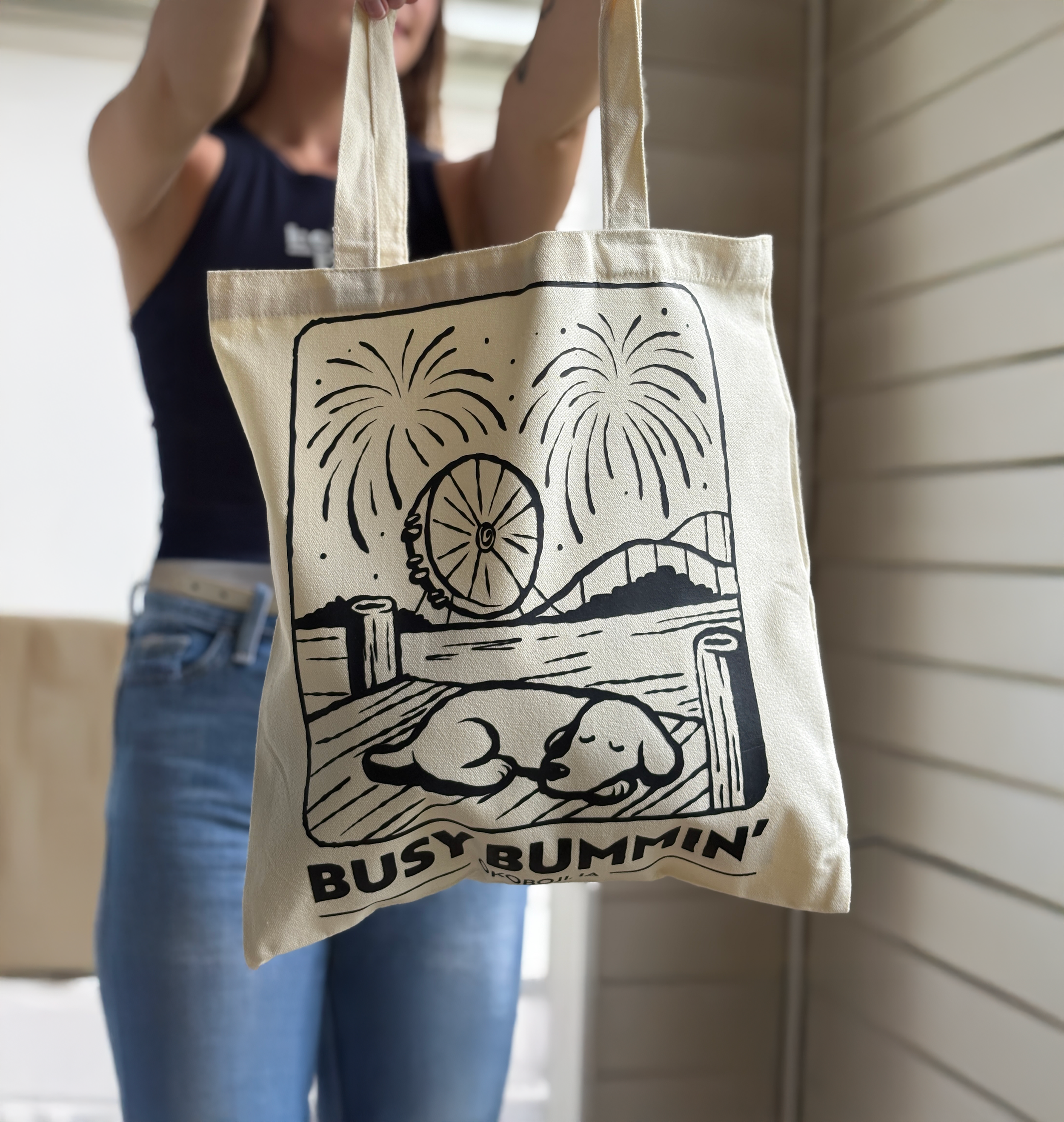 A person holding up a beige tote bag with a black illustration of fireworks, a Ferris wheel, a roller coaster, and two sleeping dogs with the text "BUSY BUMMIN'" below.