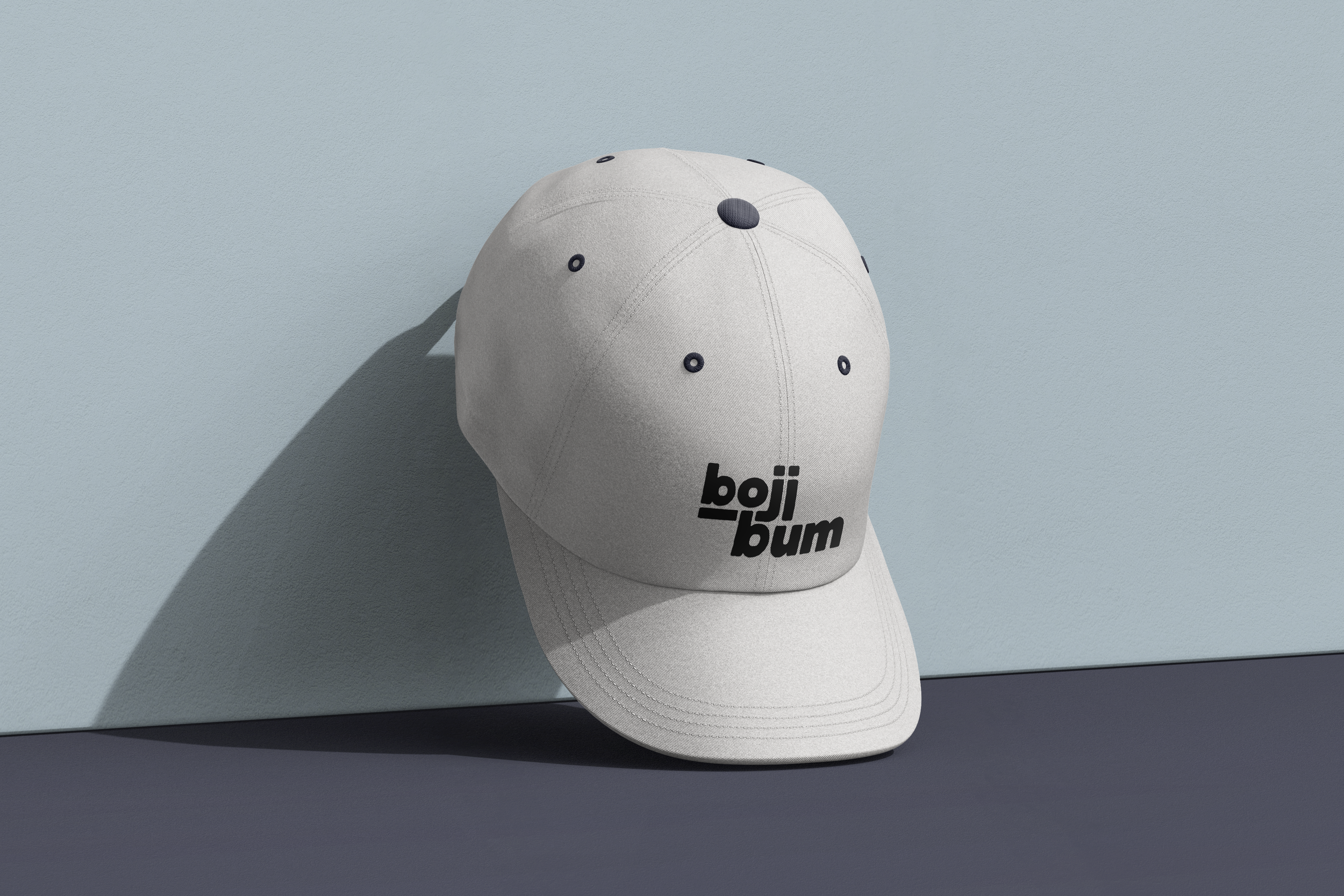 White baseball cap with black adjustable strap and logo that reads 'boji bum' in black text, placed against a light blue background with a shadow cast on the wall.