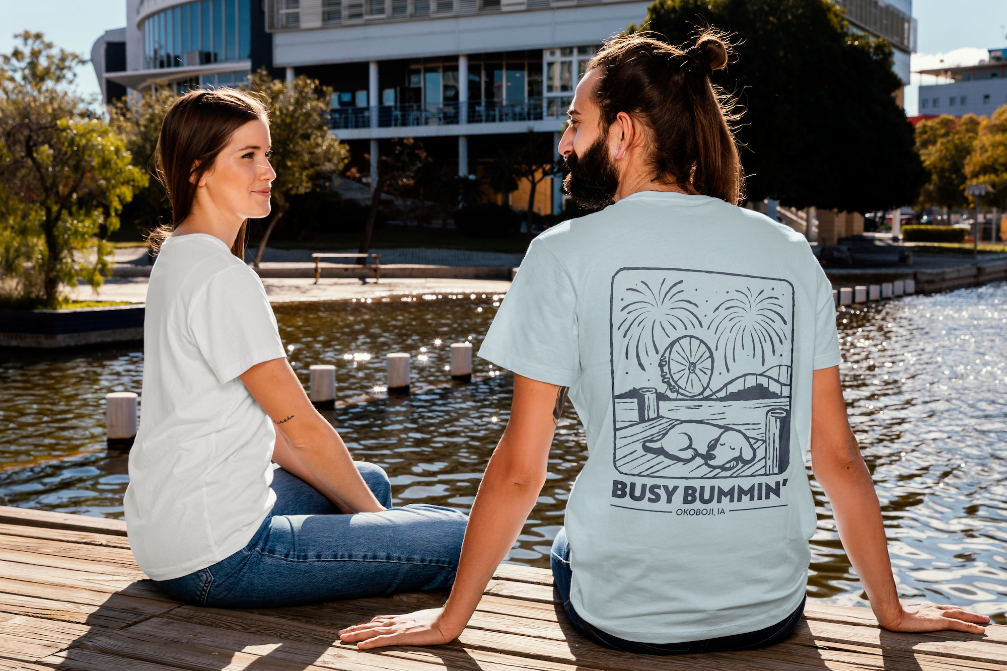 A man and woman sitting on a wooden dock by the water, talking, with buildings and trees in the background on a sunny day.