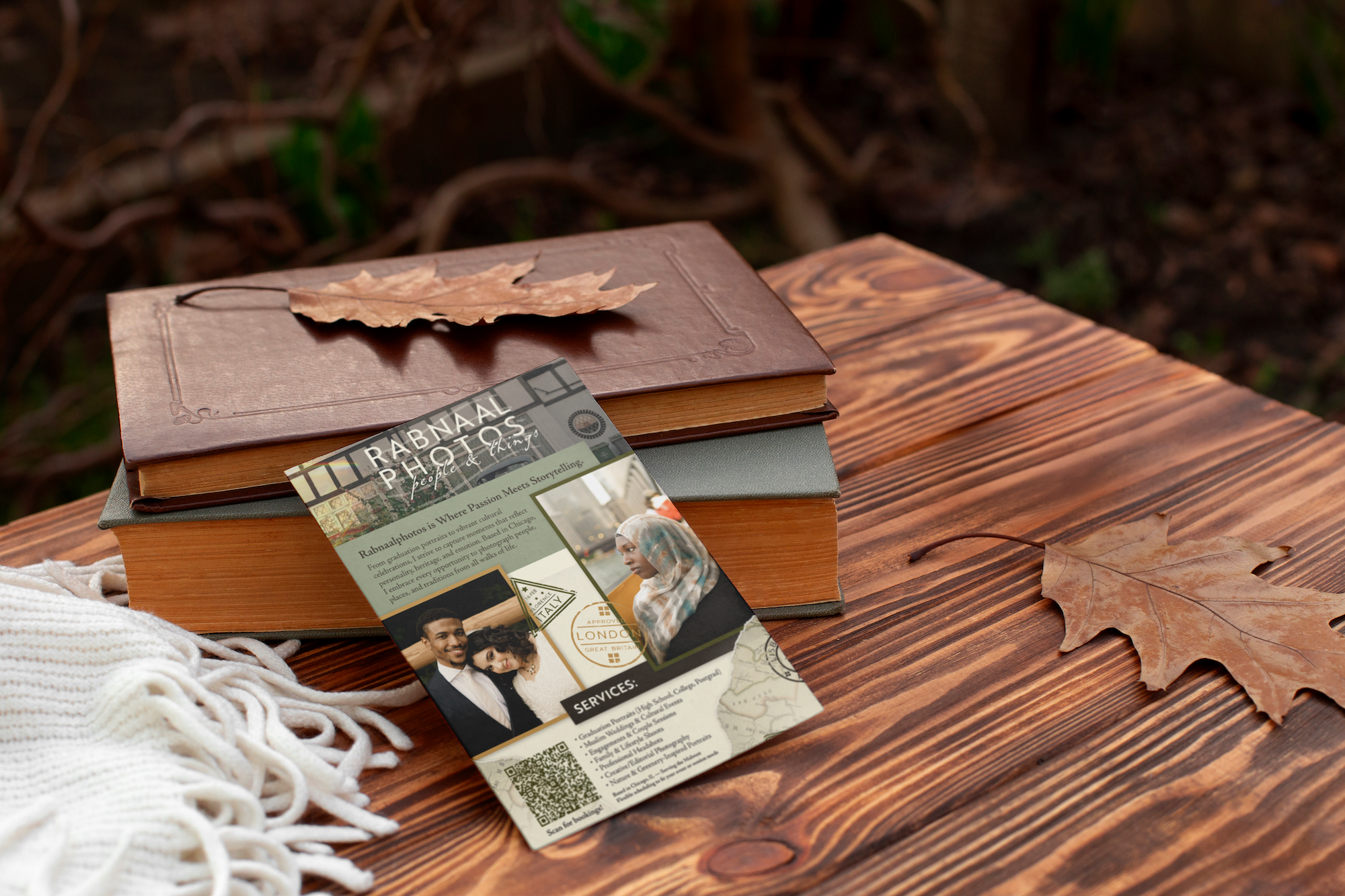 An outdoor wooden table holding a closed brown leather-bound book with a dried leaf on top, a gray notebook underneath, a leaf on the table, a white knitted cloth with fringe, and a colorful flyer with promotional content, a photo of a woman in a hij