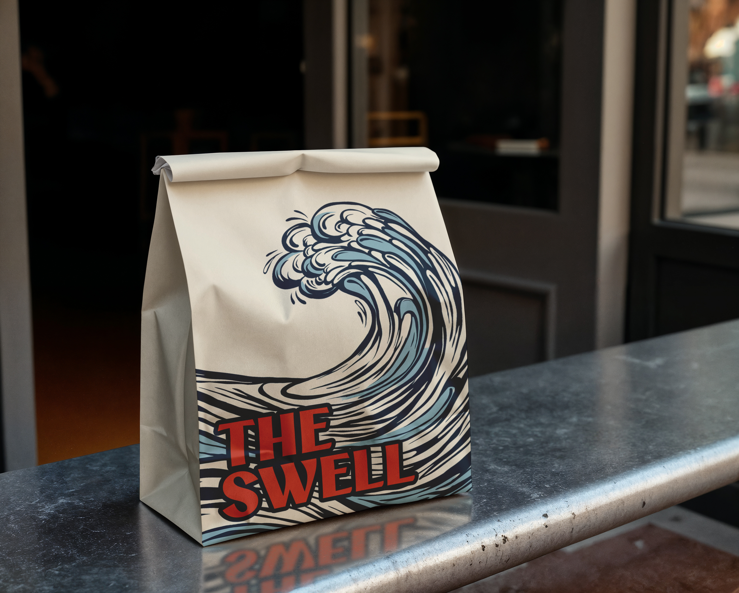 A paper bag with a graphic of a large ocean wave and the text "The Swell" in bold red letters, placed on a metal countertop.