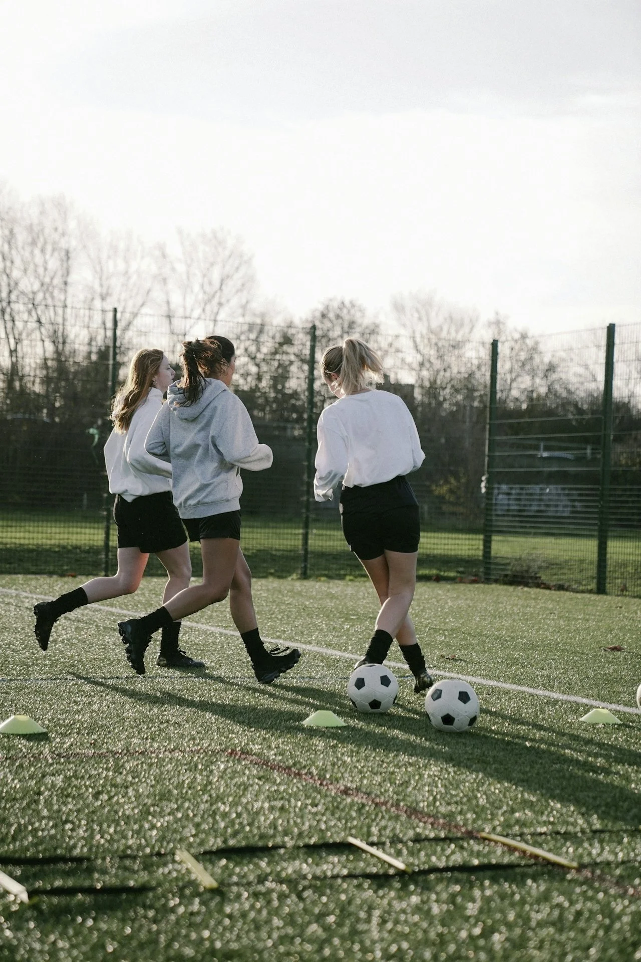 Four girls in athletic gear practicing soccer drills on a field with cones and soccer balls, during daytime with cloudy sky and trees in the background.