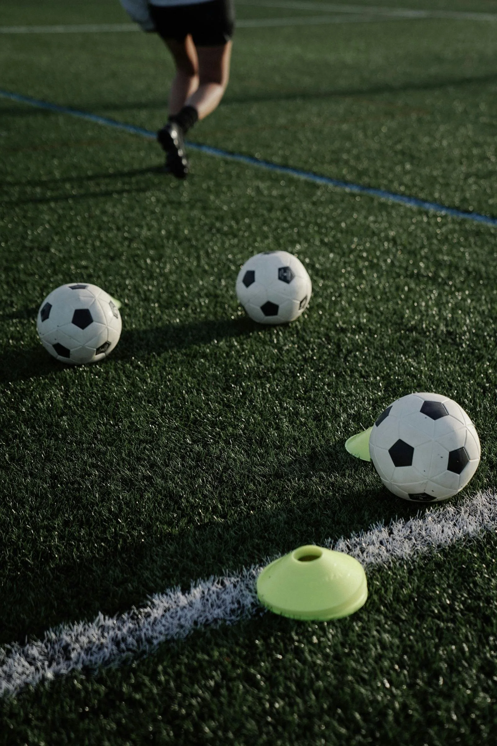 A soccer practice setup on a field with three small soccer balls and two yellow training cones near the sideline. A person's legs are visible in the background.