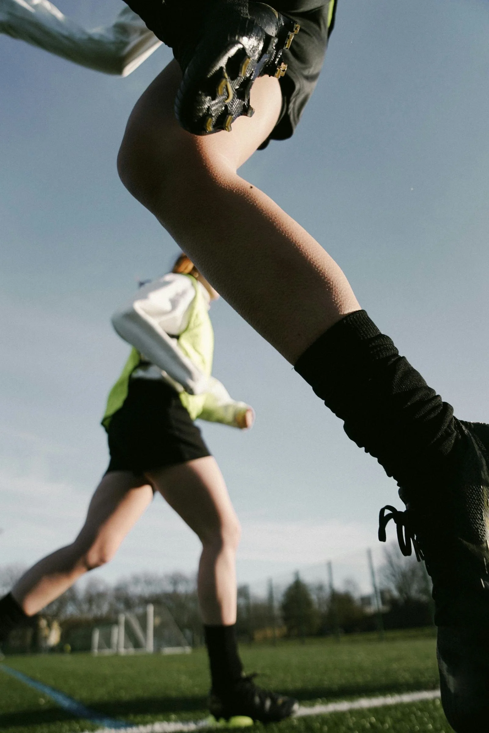 Close-up of a runner's leg in black athletic shoes and black socks, seen from below during a race or practice on a grassy field, with another runner in the background.
