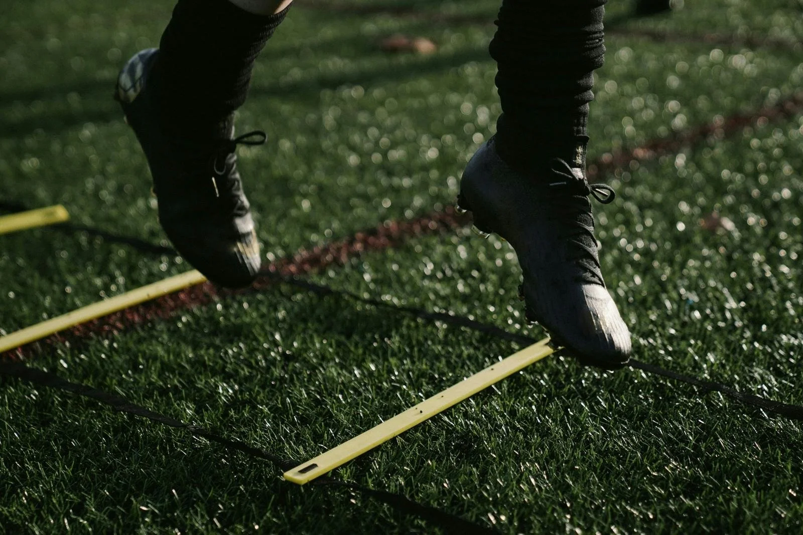 A person wearing black athletic shoes and black pants is practicing agility drills on a grassy field. They are stepping over yellow and black agility ladders laid flat on the ground.