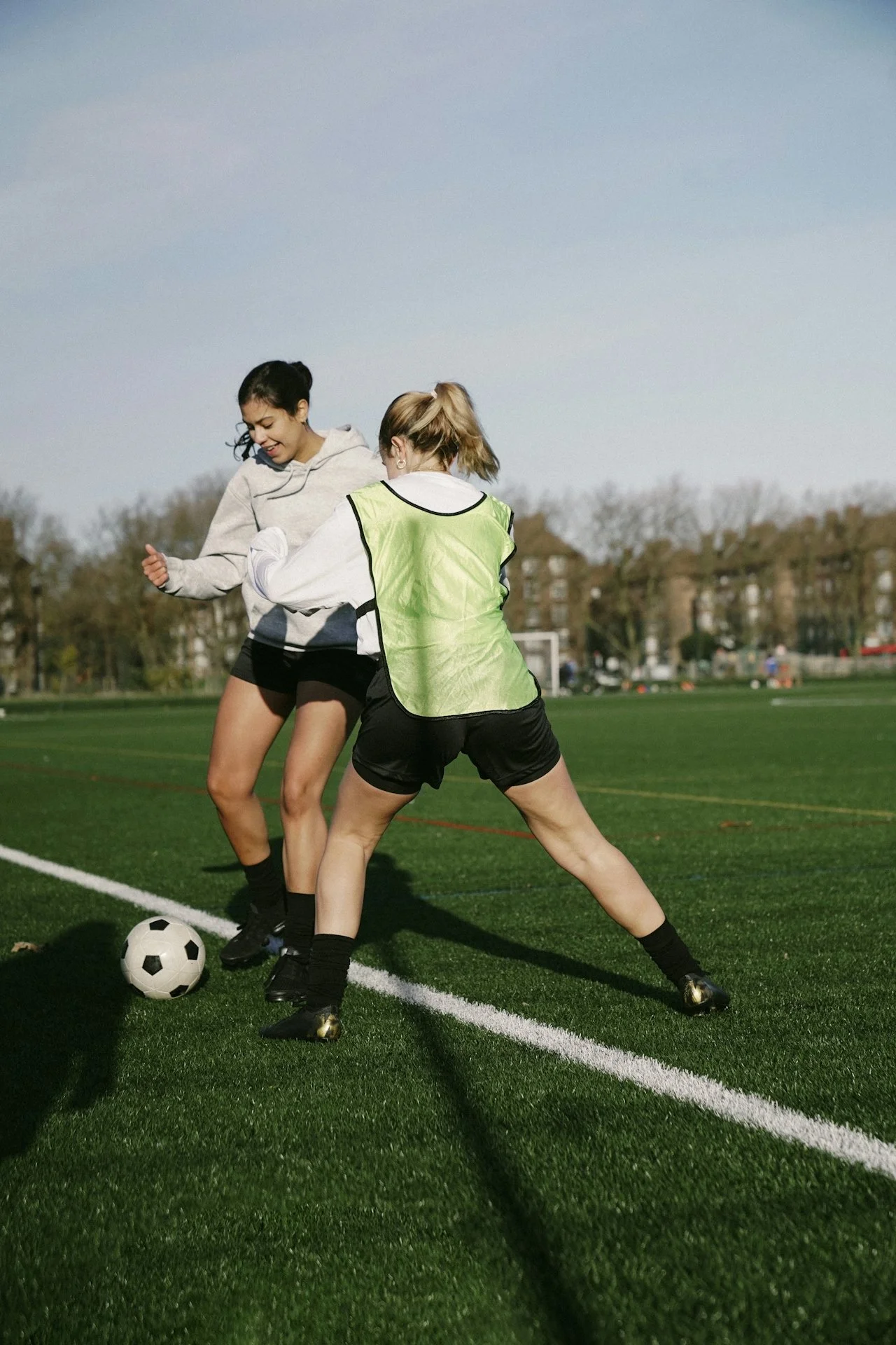 Two women playing soccer on a field, one in a gray hoodie and shorts, the other in a yellow vest, actively contesting for the ball on a green artificial turf field with trees and buildings in the background.