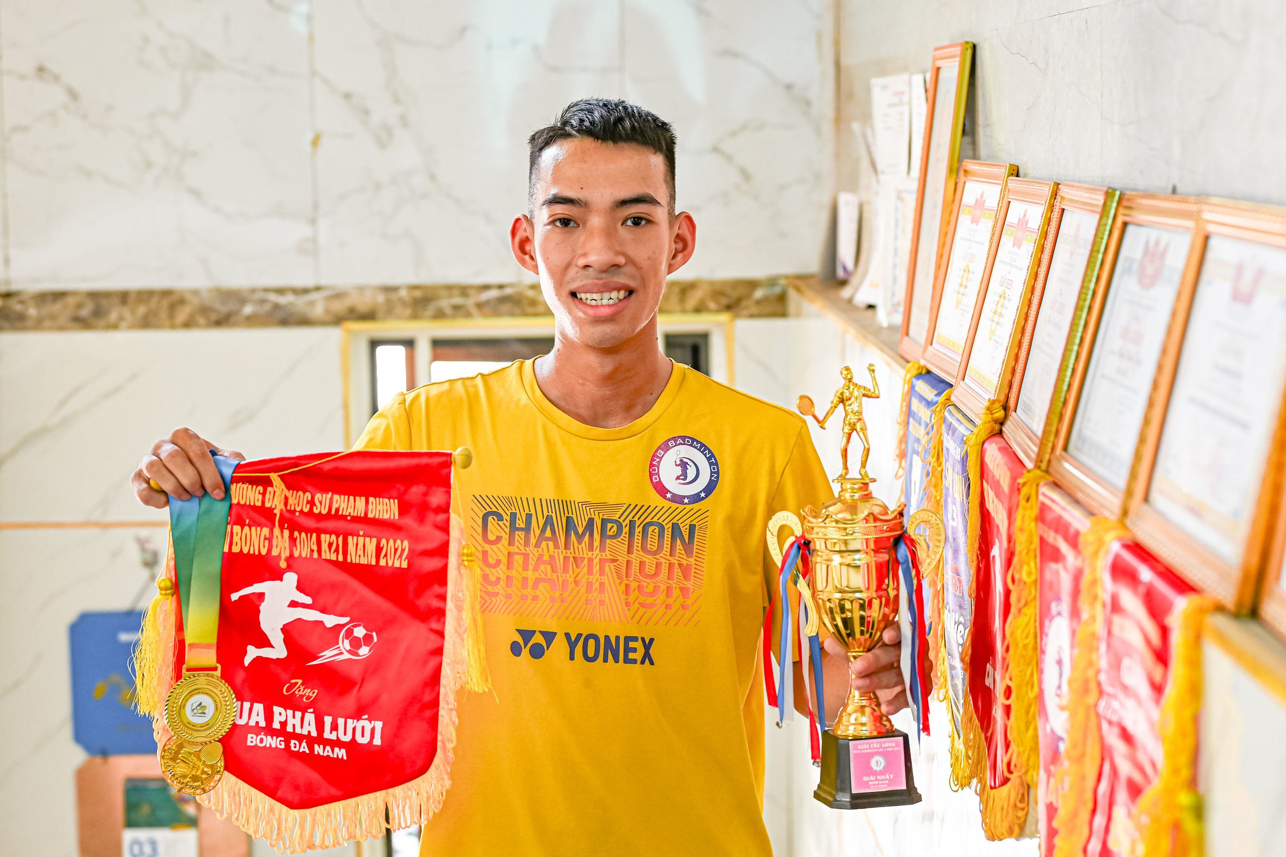 A young man wearing a yellow sports shirt standing indoors, holding medals, a red flag, and a trophy, with medals and banners displayed on a wall behind him.
