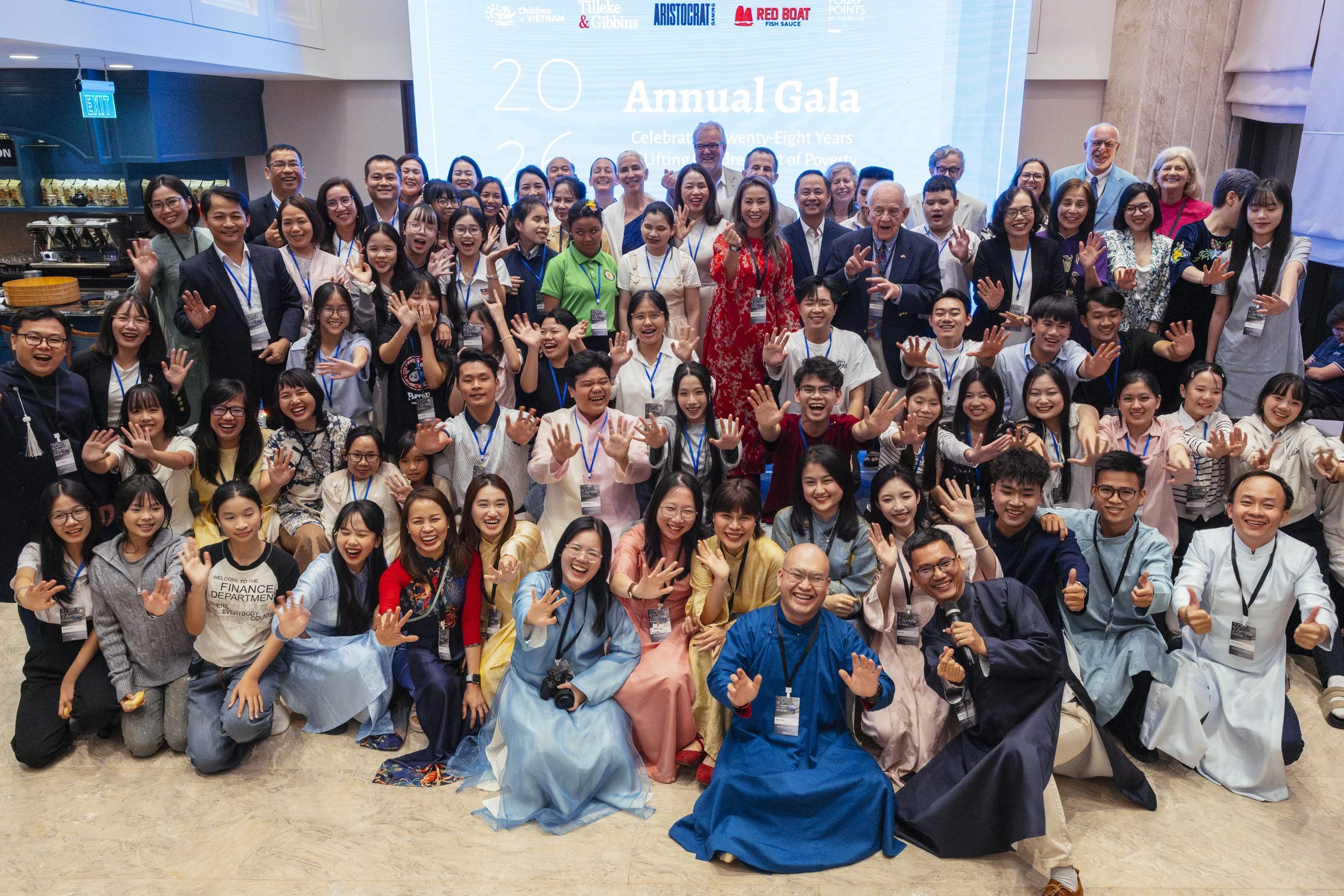 A large group of people, including children and adults, smiling and waving at the camera during COV's 26th Annual Gala, with a blue screen behind them displaying the event details.