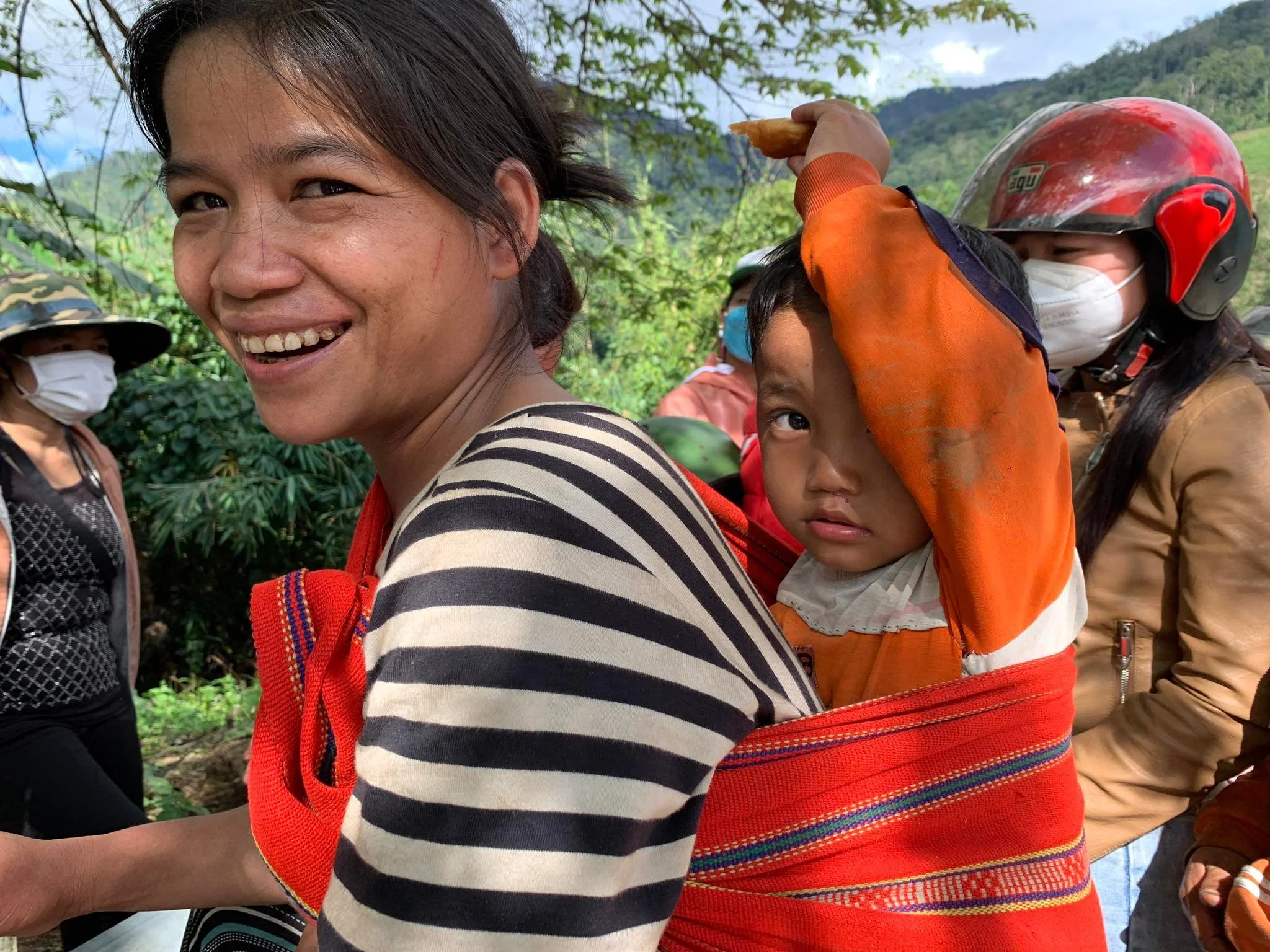 A Hmong woman smiling while carrying her child on her back, with green trees and hills in the background.