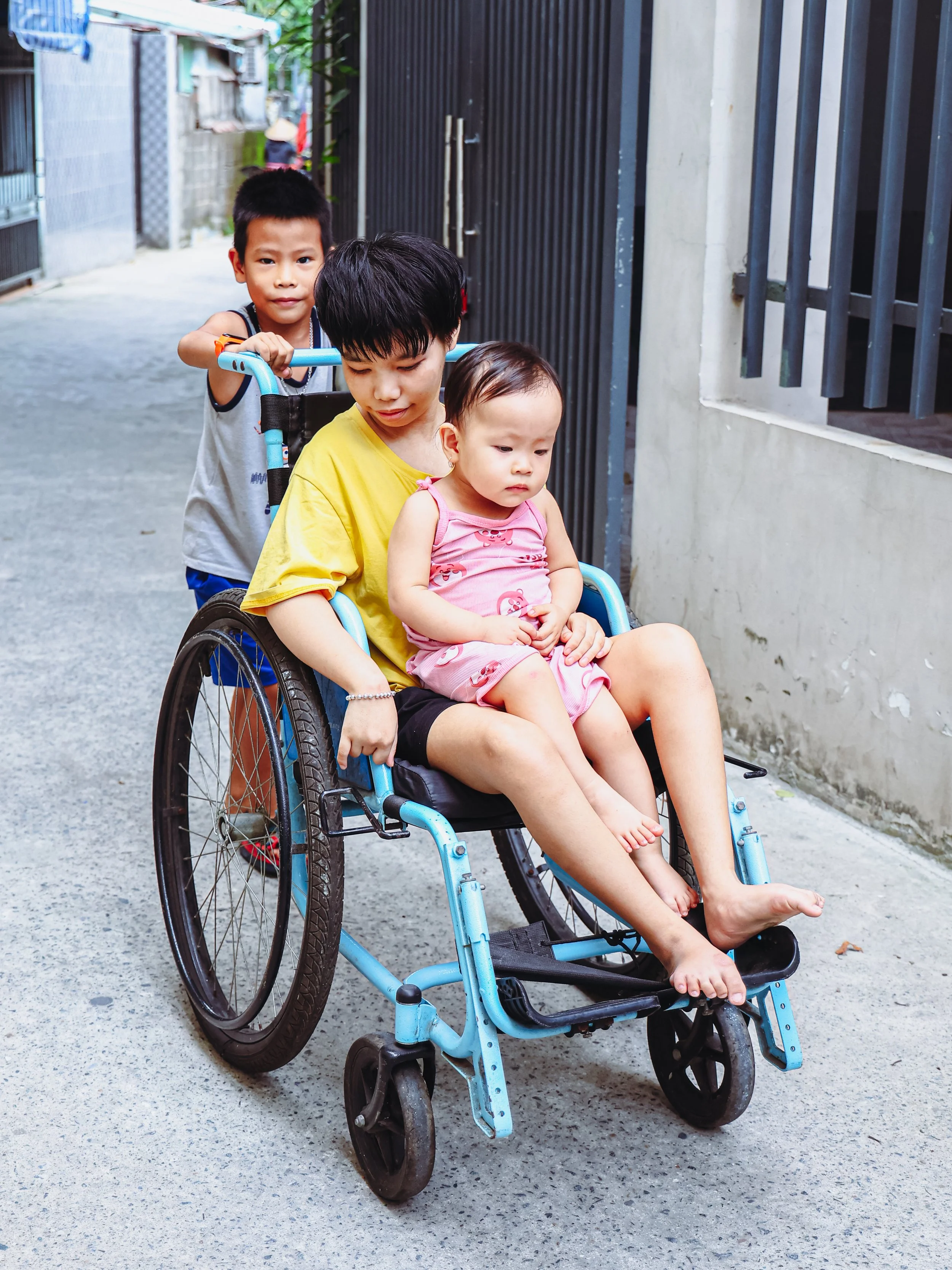 A woman in a wheelchair with a young girl in a pink dress sitting on her lap, and a young boy pushing the wheelchair from behind on a narrow street with residential buildings.