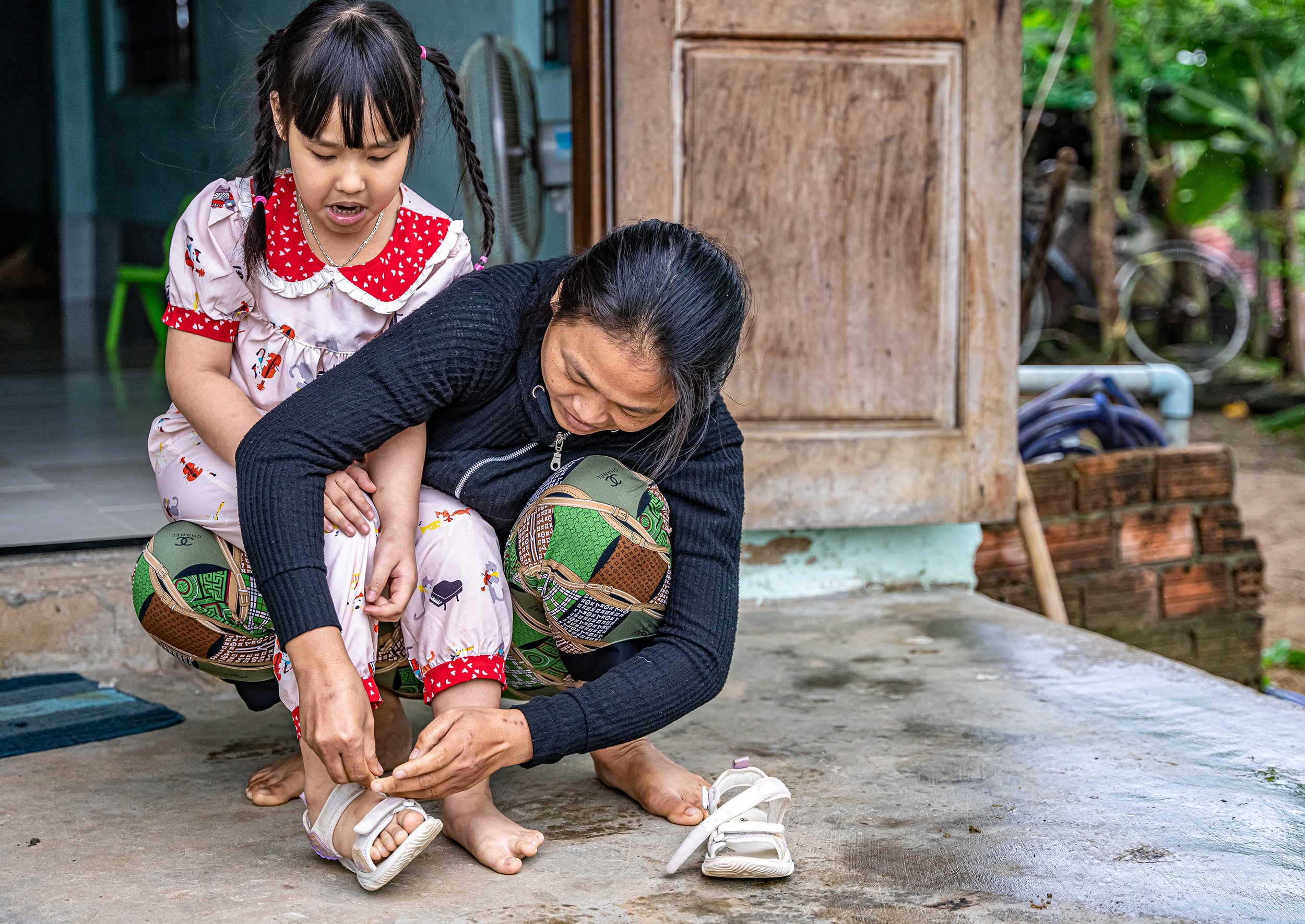 A young girl with braided hair and a woman, both barefoot, are tying their shoes outside near a wooden structure. The girl is wearing a patterned dress, and the woman is in a black jacket. Green plants are in the background.