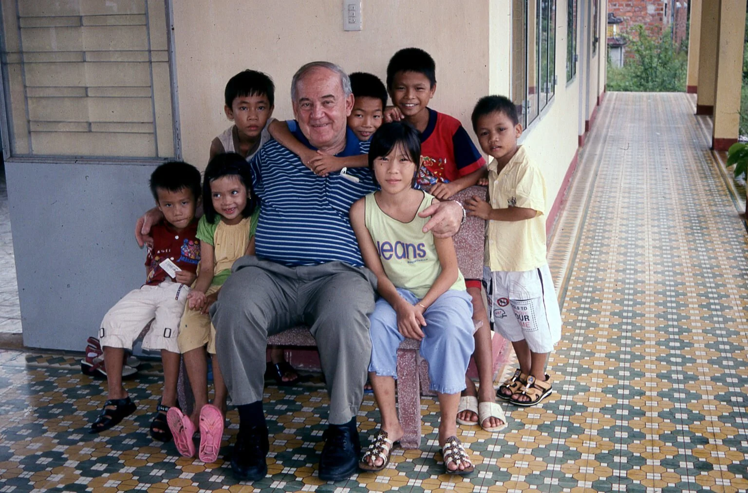 COV founder Benjamin Wilson, surrounded by children, sitting on a bench in a corridor with a decorated tile floor and yellow walls.