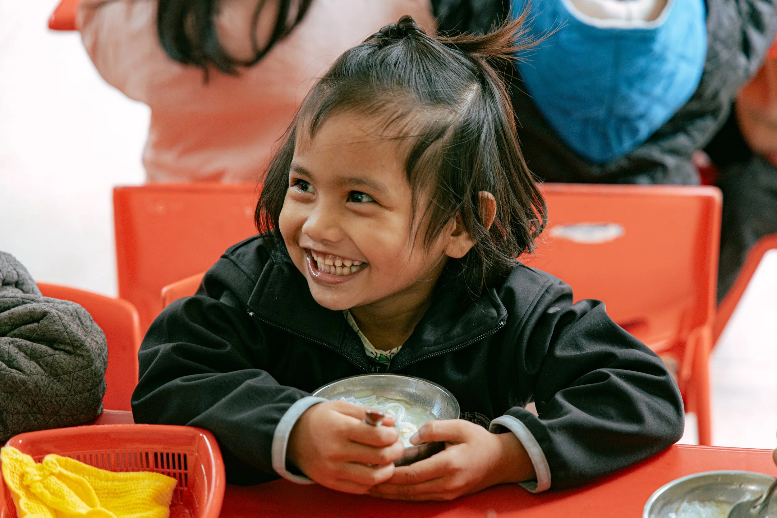 A young girl with dark hair and bright eyes smiling while eating soup at a table in a cafeteria, with orange chairs and other people blurred in the background.