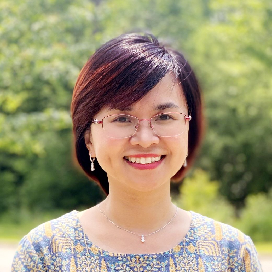 A woman with short dark hair and glasses, smiling outdoors with greenery in the background.