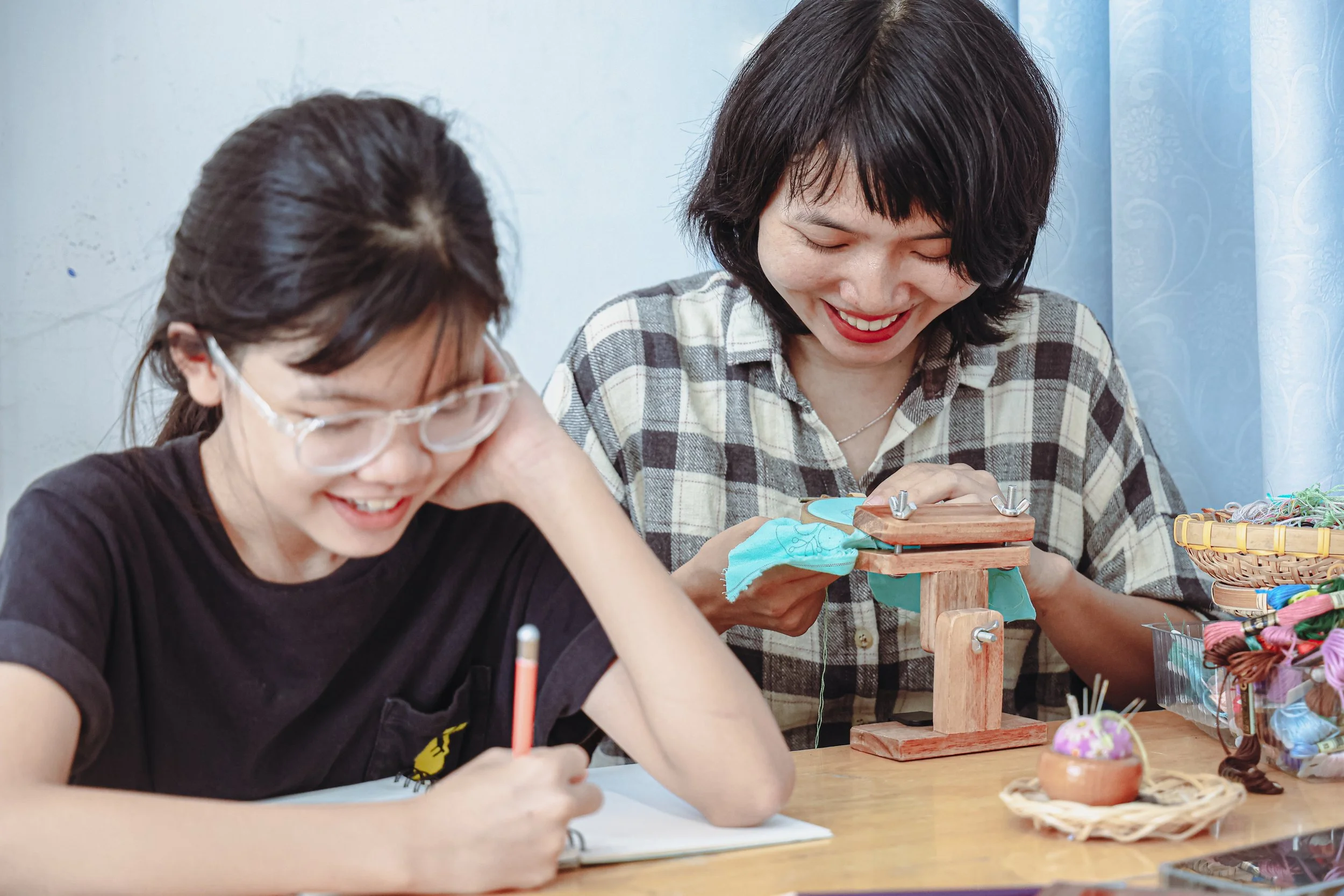 Two women smiling and working on a craft project at a table with colorful threads, fabric, and small decorative items, surrounded by a variety of craft supplies.