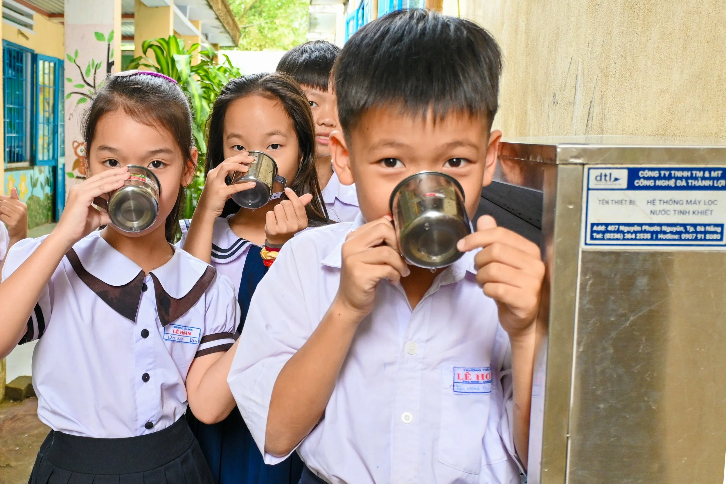 Children in school uniforms drinking water from stainless steel cups at an outdoor water fountain.