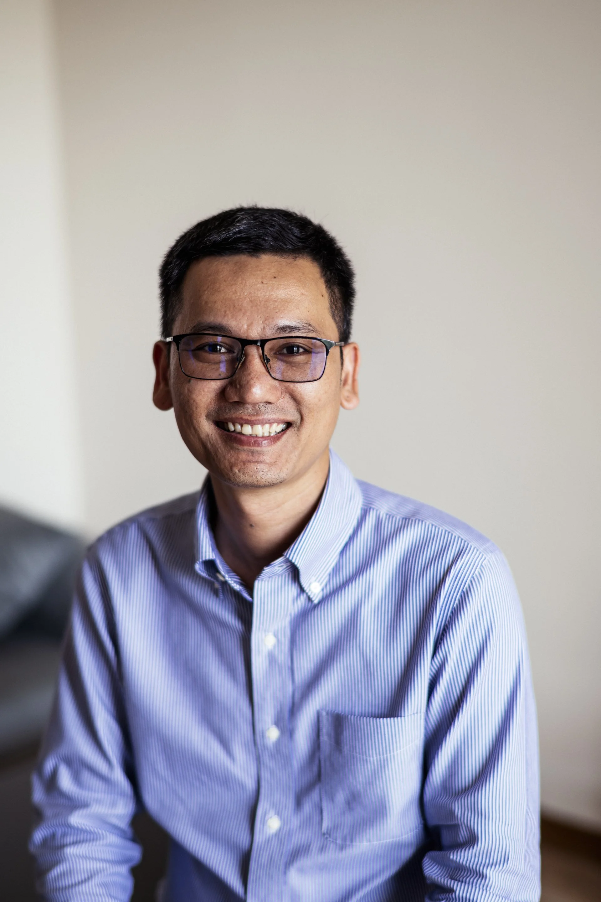 A smiling man with short dark hair, glasses, wearing a blue and white striped button-up shirt, sitting in a well-lit room with a plain background.