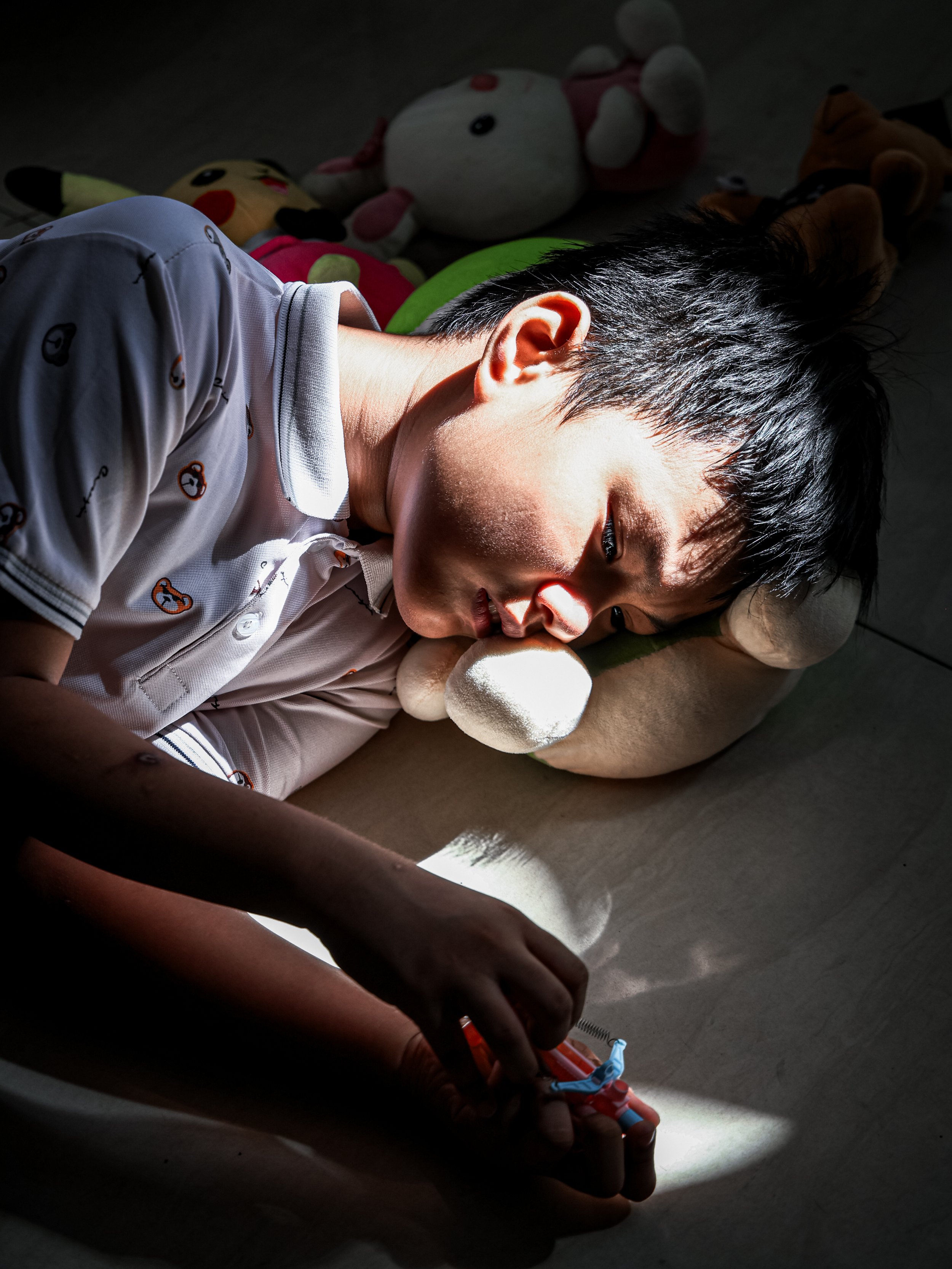 A young boy lying on the floor, asleep, with sunlight shining on his face and head. There are stuffed animals around him, including Pikachu and Hello Kitty, with some of them visible in the background.