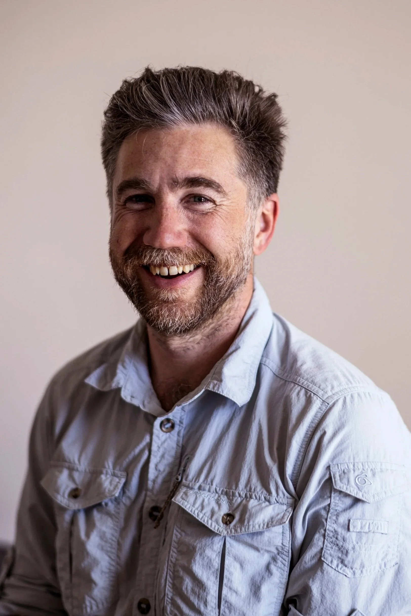 A smiling man with a beard and styled hair wearing a light-colored button-up shirt, sitting against a plain beige background.