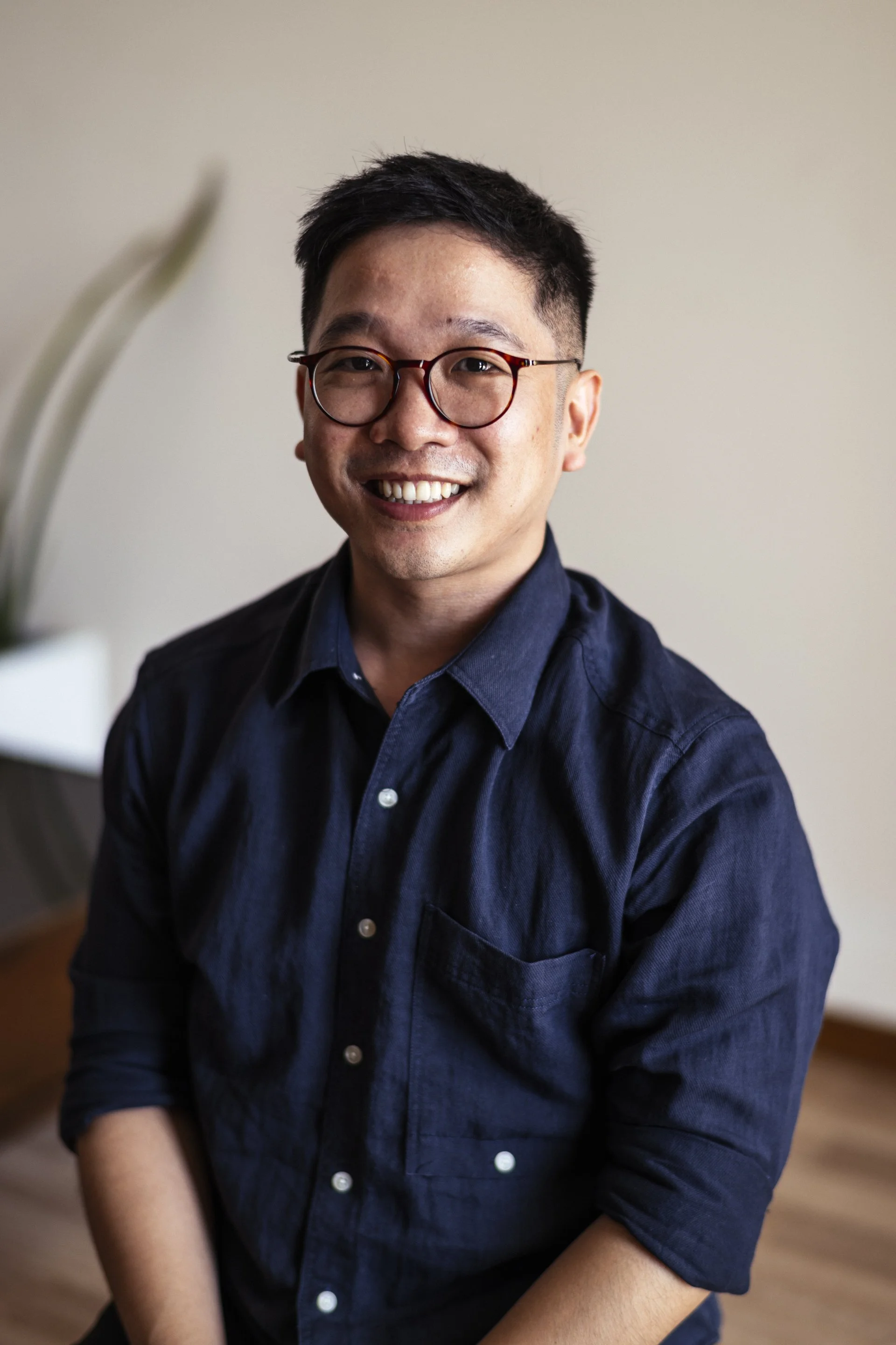 A smiling man with short dark hair, glasses, and wearing a navy blue button-up shirt, seated indoors against a beige wall.