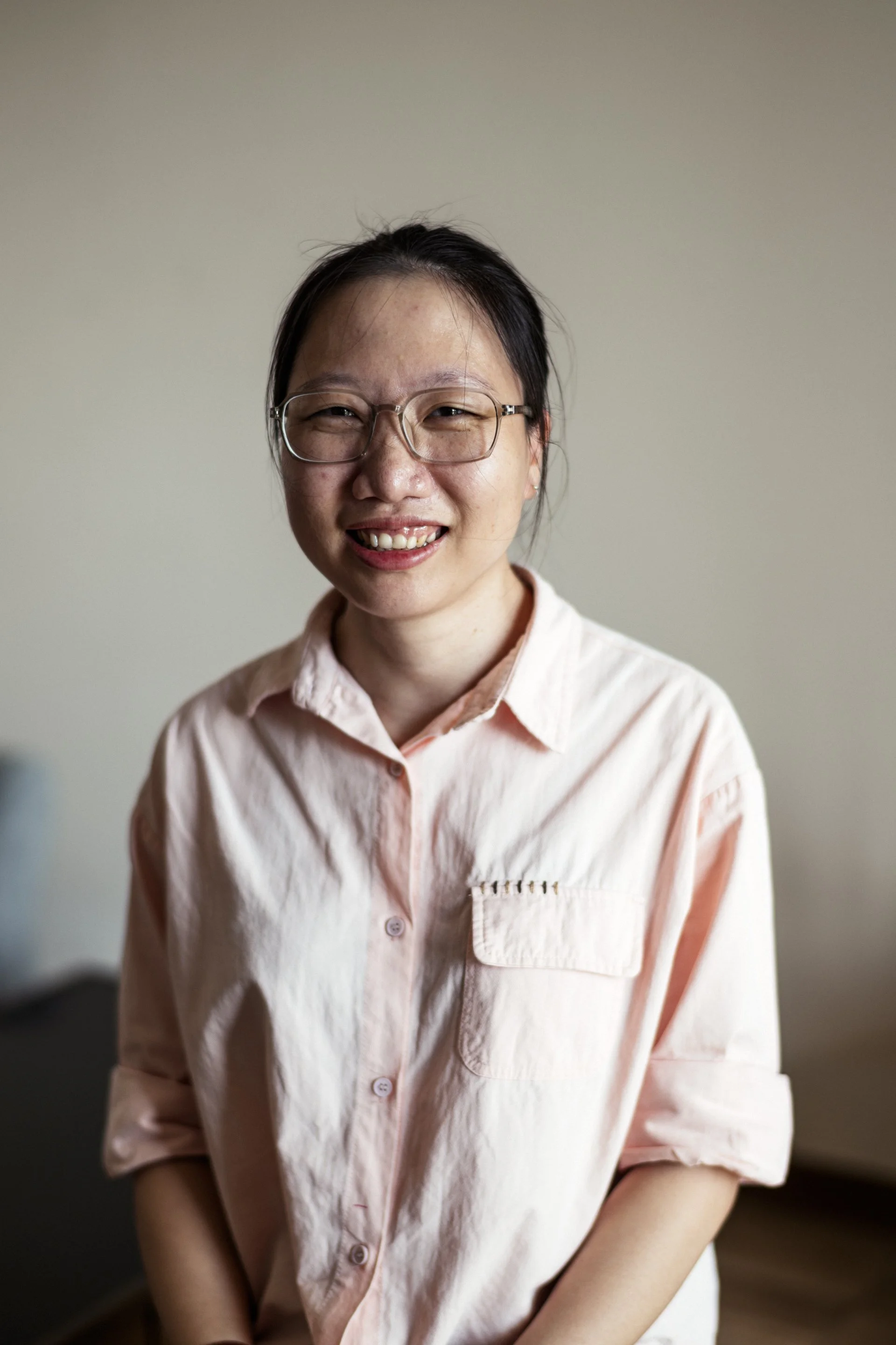 A woman with black hair, glasses, and a light pink button-up shirt smiling at the camera against a neutral wall background.