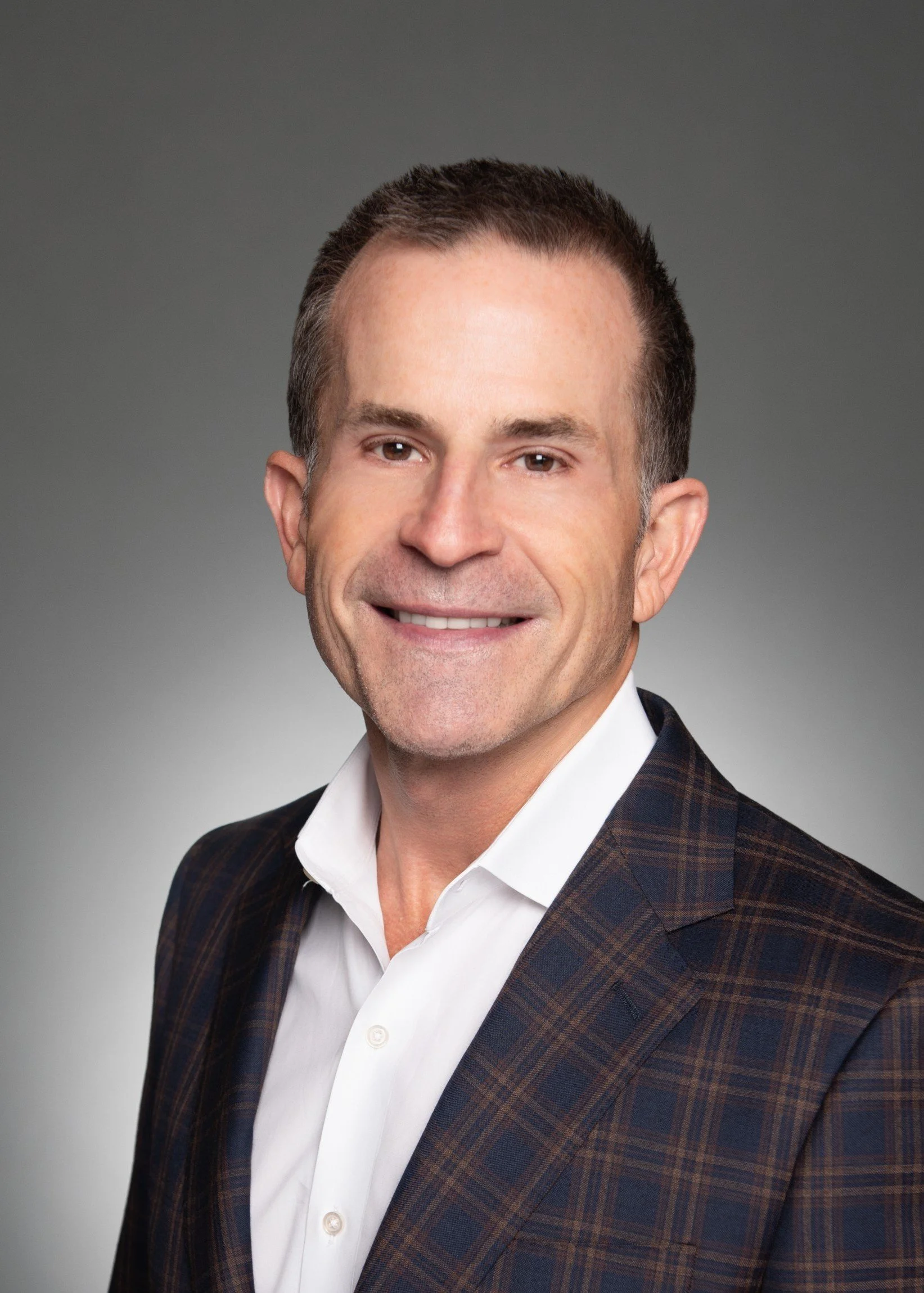 Close-up portrait of a man in a dark plaid suit with a white shirt, smiling against a plain gray background.
