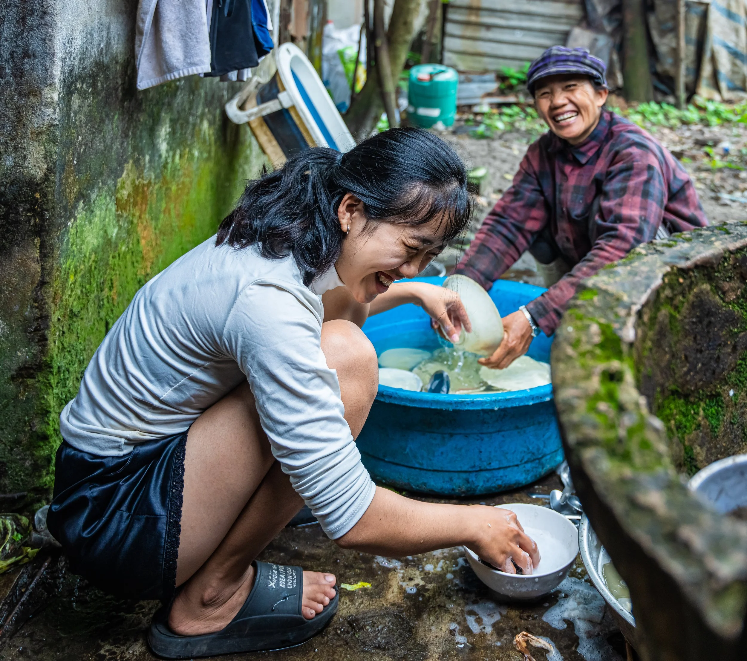 Two women are crouched on the ground outdoors, laughing and washing clothes in a blue basin filled with water. There is green moss on a wall behind them, and clothes hanging to dry above. One woman is wearing a white shirt, and the other is wearing a plaid shirt with a hat. A green container and some rocks are also visible.