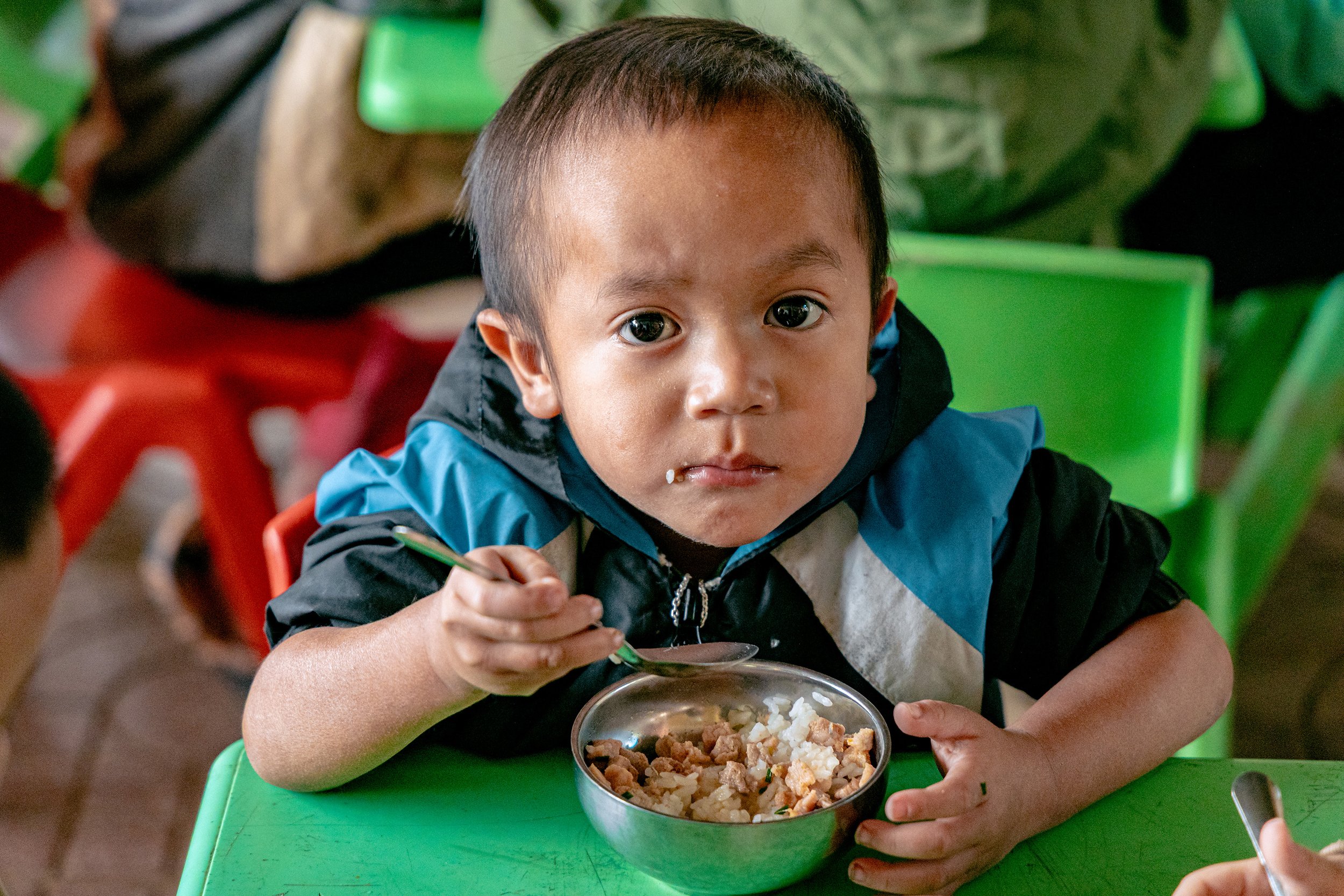 A young boy with short dark hair and big eyes sitting at a green table, holding a fork and eating food from a metal bowl, with rice and meat. He is wearing a blue and black jacket, and other children are partially visible around him.