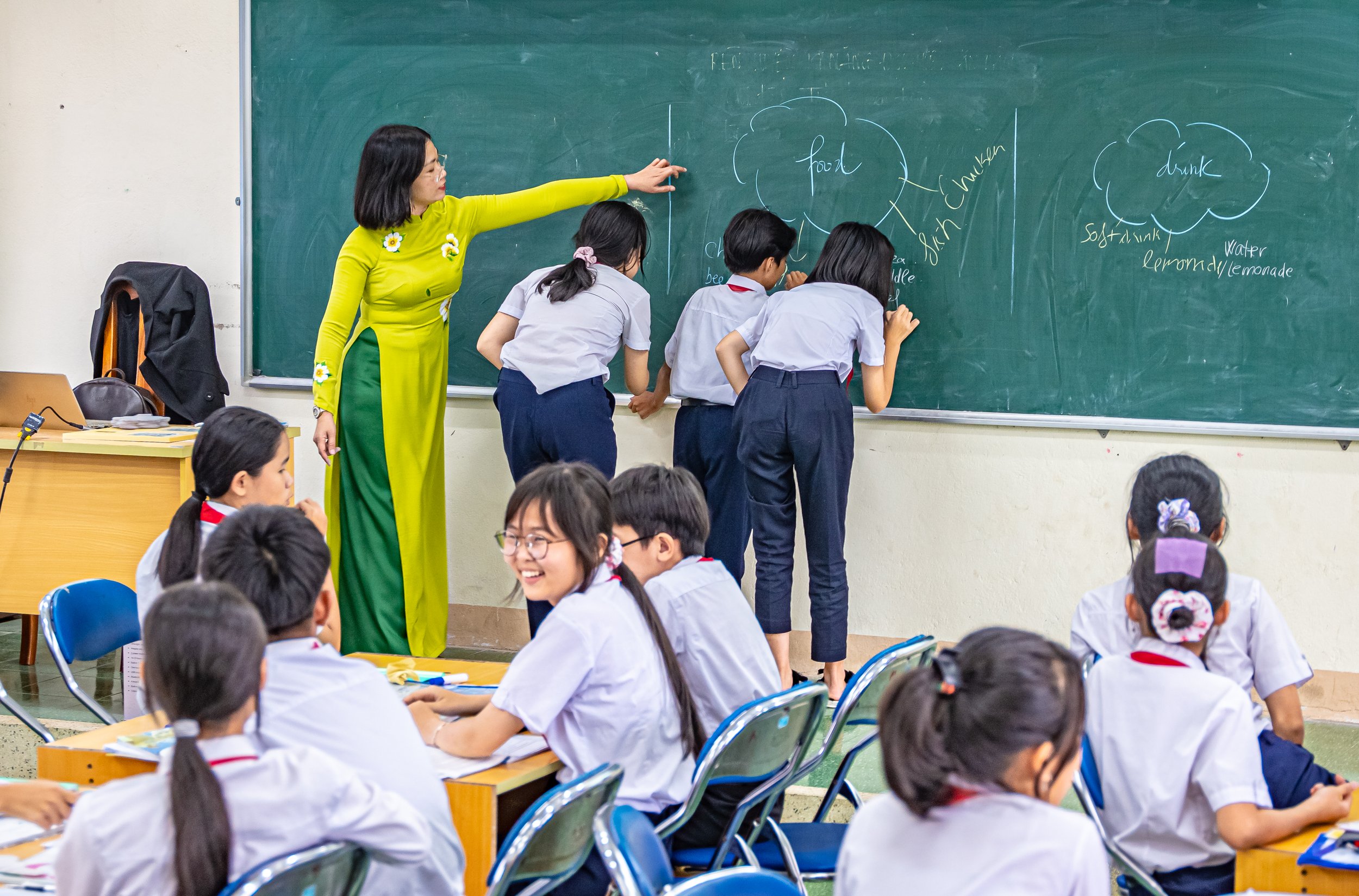 A classroom with students in white uniforms seated at desks, a teacher in a yellow and green dress pointing to a blackboard, which has drawings of clouds labeled "food," "drink," and "soft drink/lemonade," and other notes.
